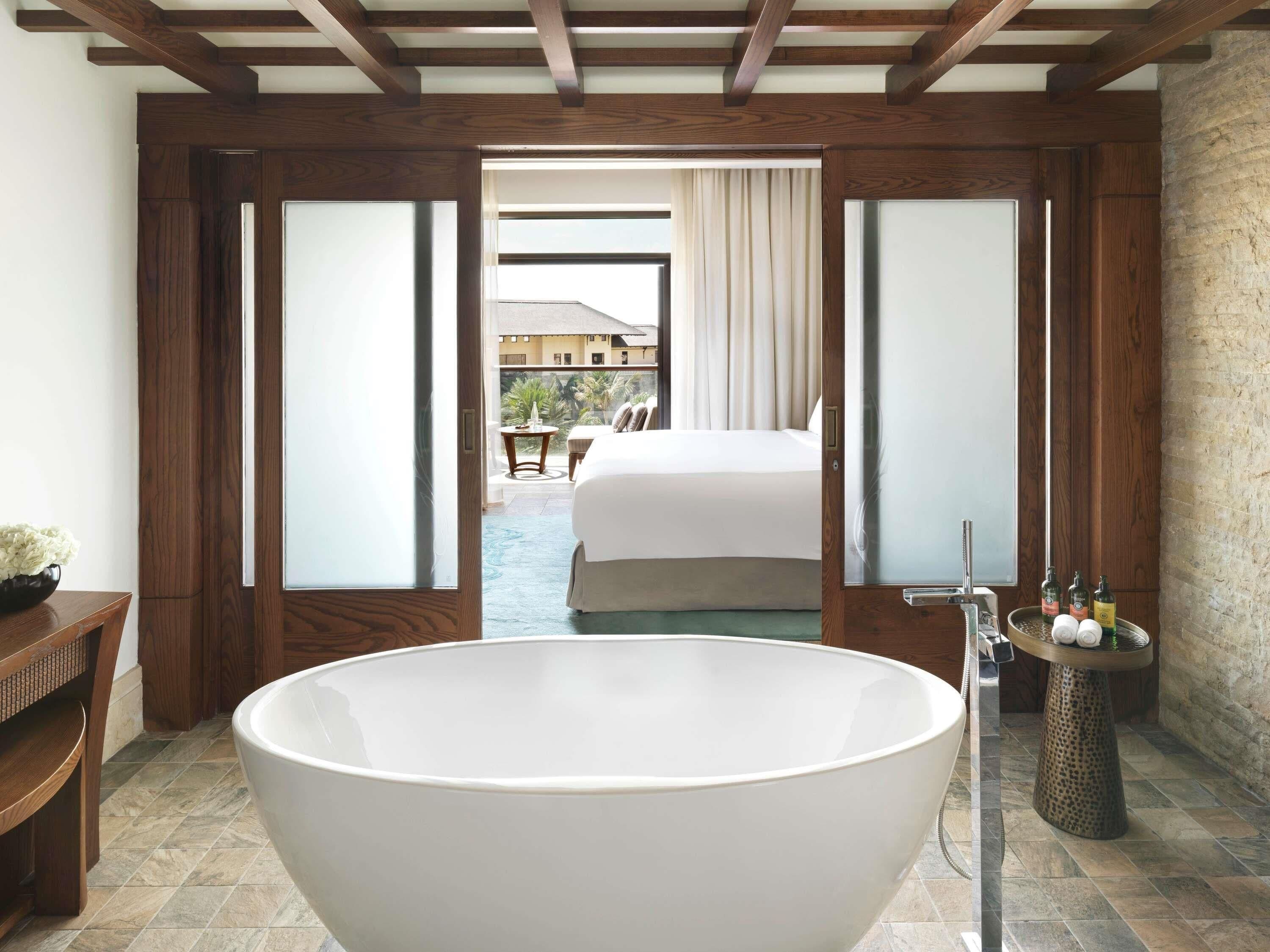 A modern bathroom with a large freestanding bathtub, wooden flooring, and a view of a bedroom with a bed and curtains.