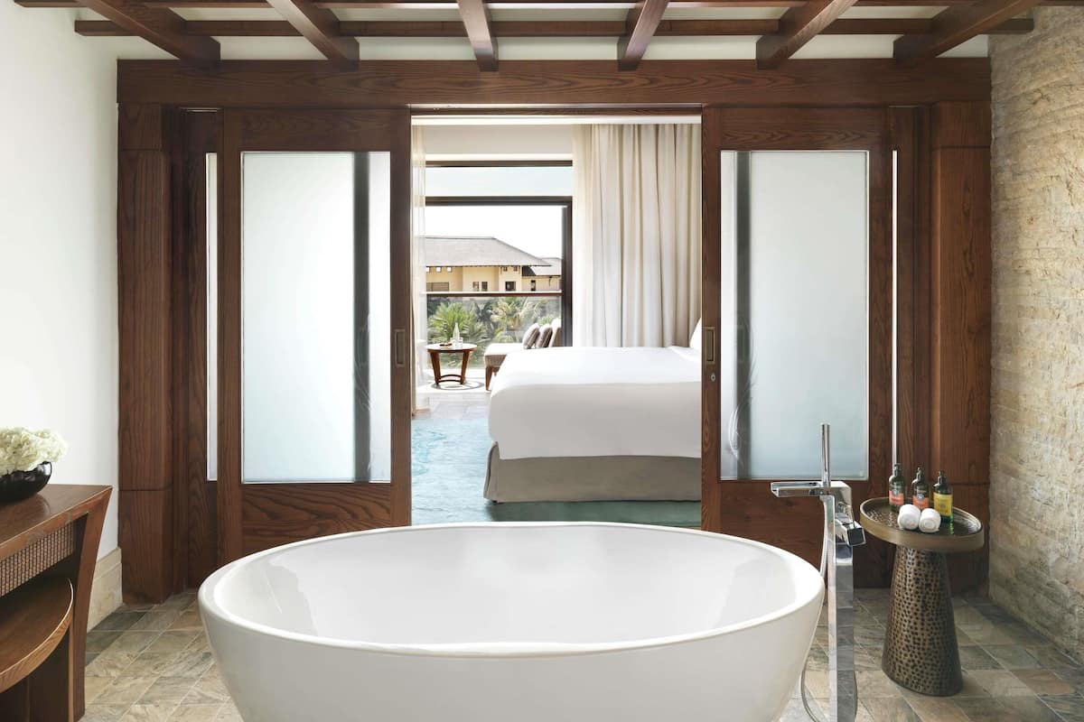 Een moderne badkamer met een ruim vrijstaand bad, houten vloeren en uitzicht op een slaapkamer met een bed en gordijnen.