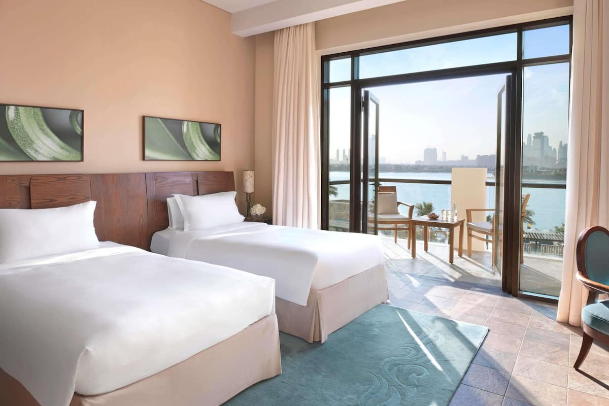 Een hotelkamer met twee bedden, een groot raam met uitzicht op een stadsbeeld en water, en een balkon met zitplaatsen.