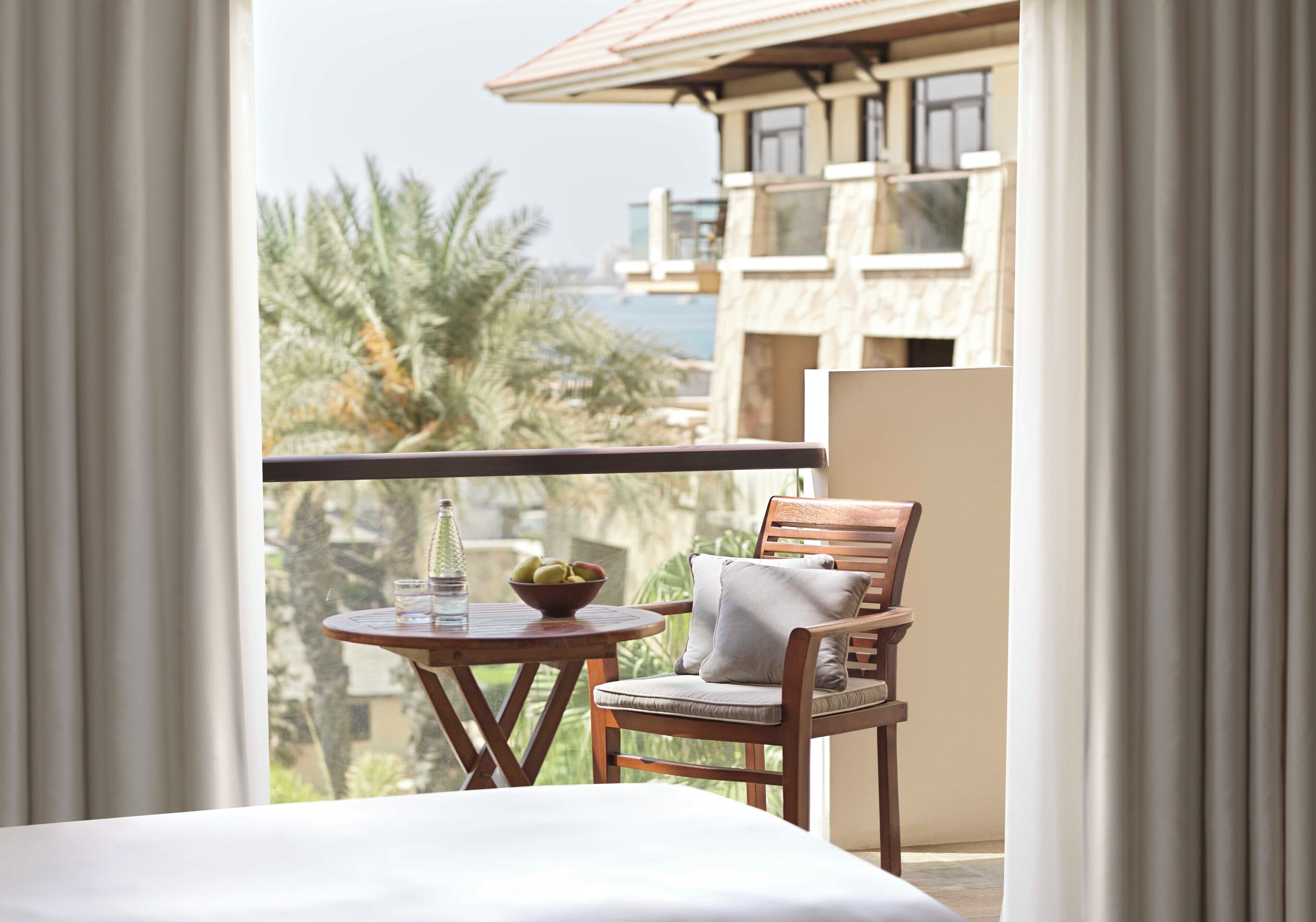 A balcony with a wooden chair, a small table with a bowl of fruit and glasses, and a view of a building and palm trees.