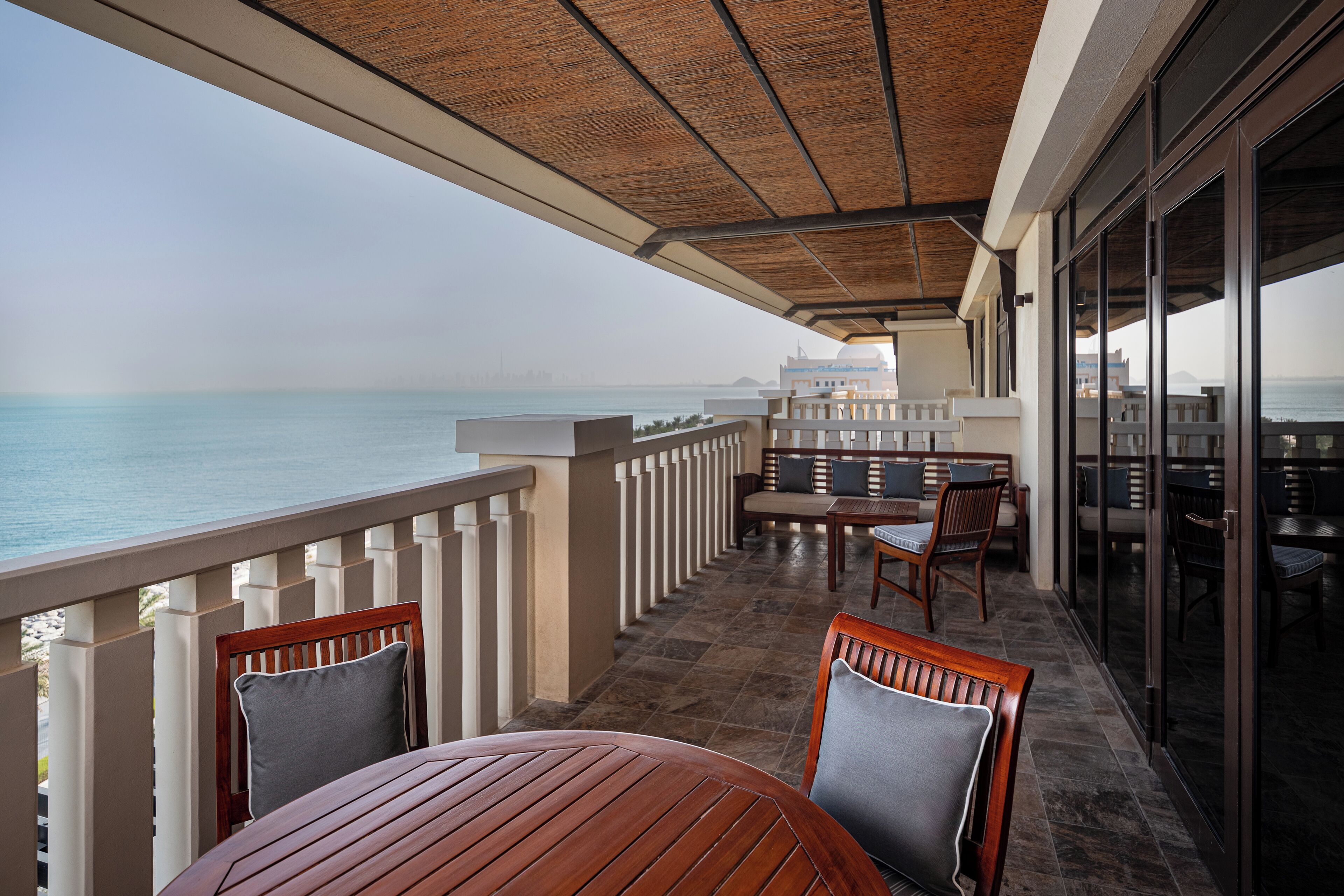 Un espace extérieur abrité avec des places assises, offrant une vue sur la mer, doté de mobilier en bois et d’une porte en verre donnant accès à un autre espace.