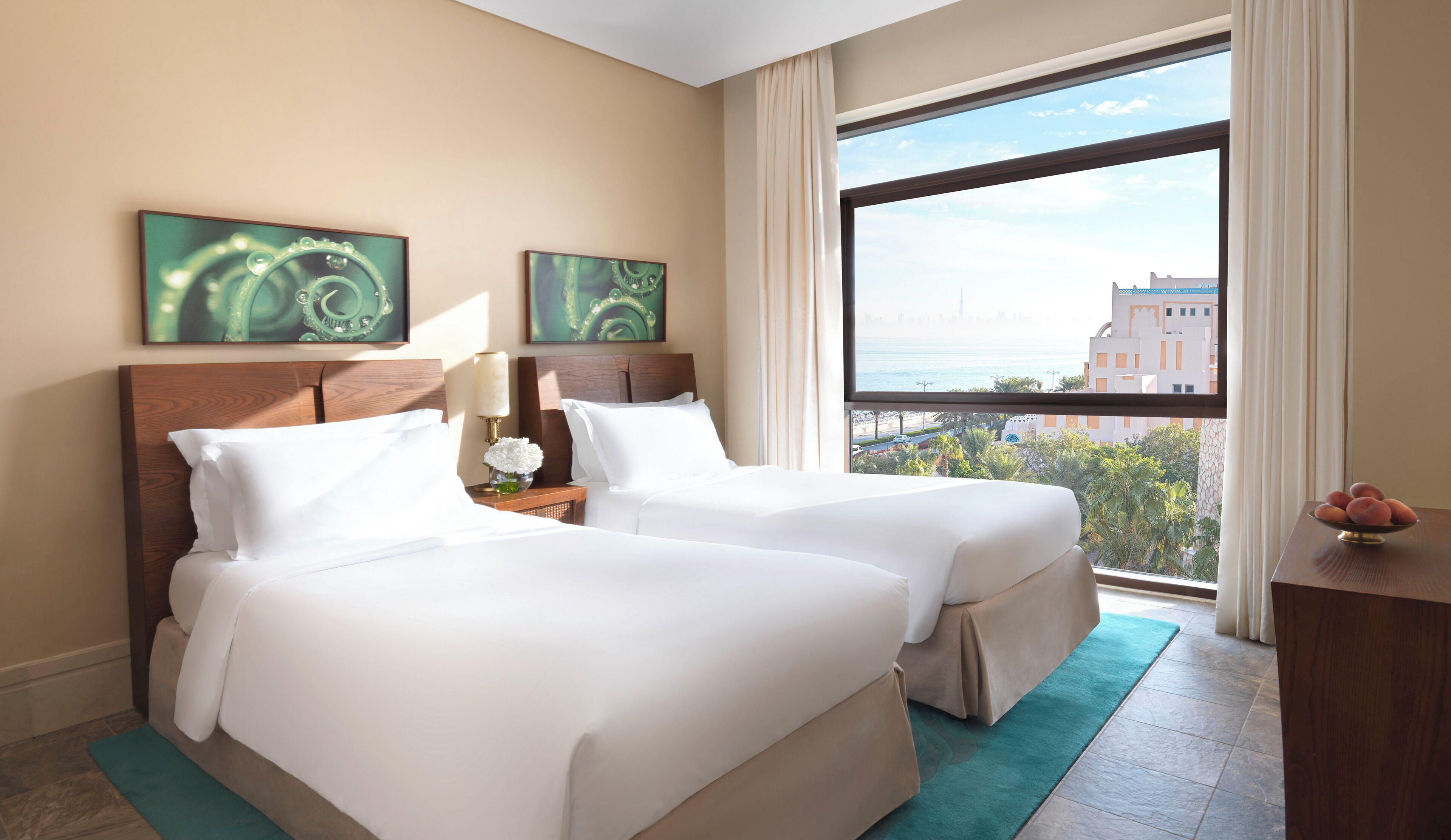 A hotel room with two beds, a large window with a city view, and artwork on the wall.