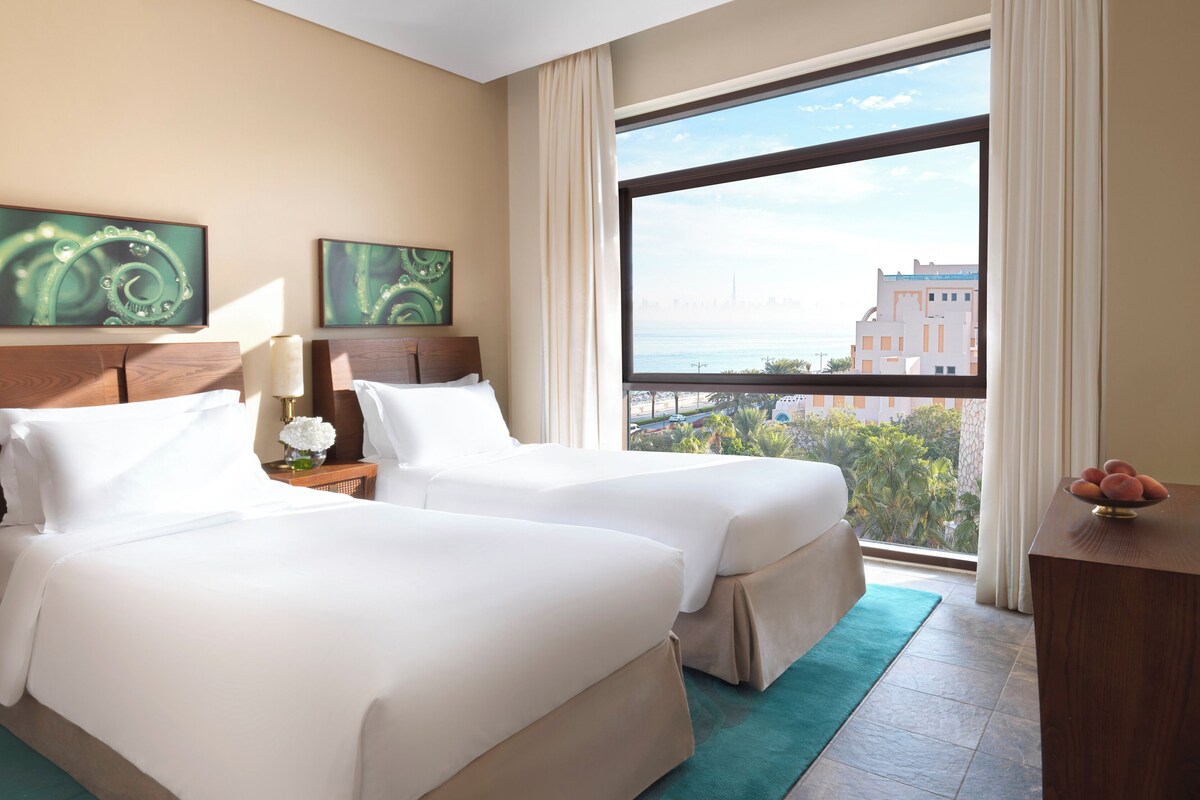 Una habitación de hotel con dos camas, un ventanal con vista a la ciudad y obras de arte en la pared.