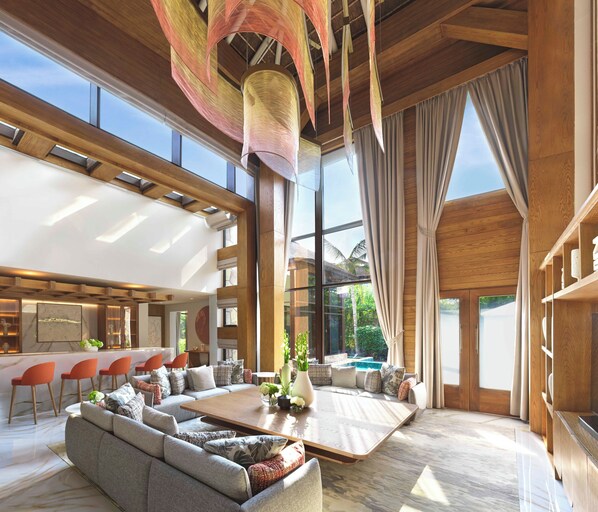 A spacious living area with a large sofa, a coffee table, and a dining area with bar stools. There is a large window with curtains and a view of greenery outside.