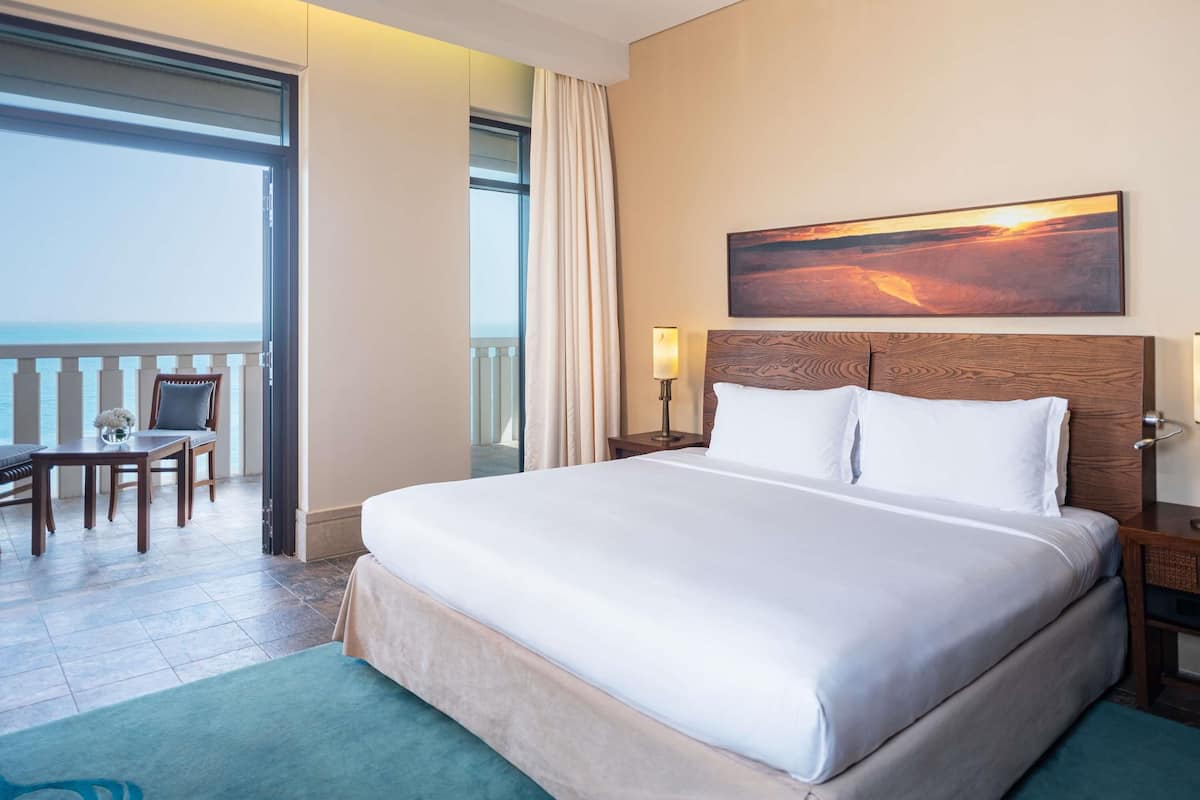 Een hotelkamer met een groot bed, een bureau met stoel, uitzicht op de oceaan en een schilderij aan de muur.