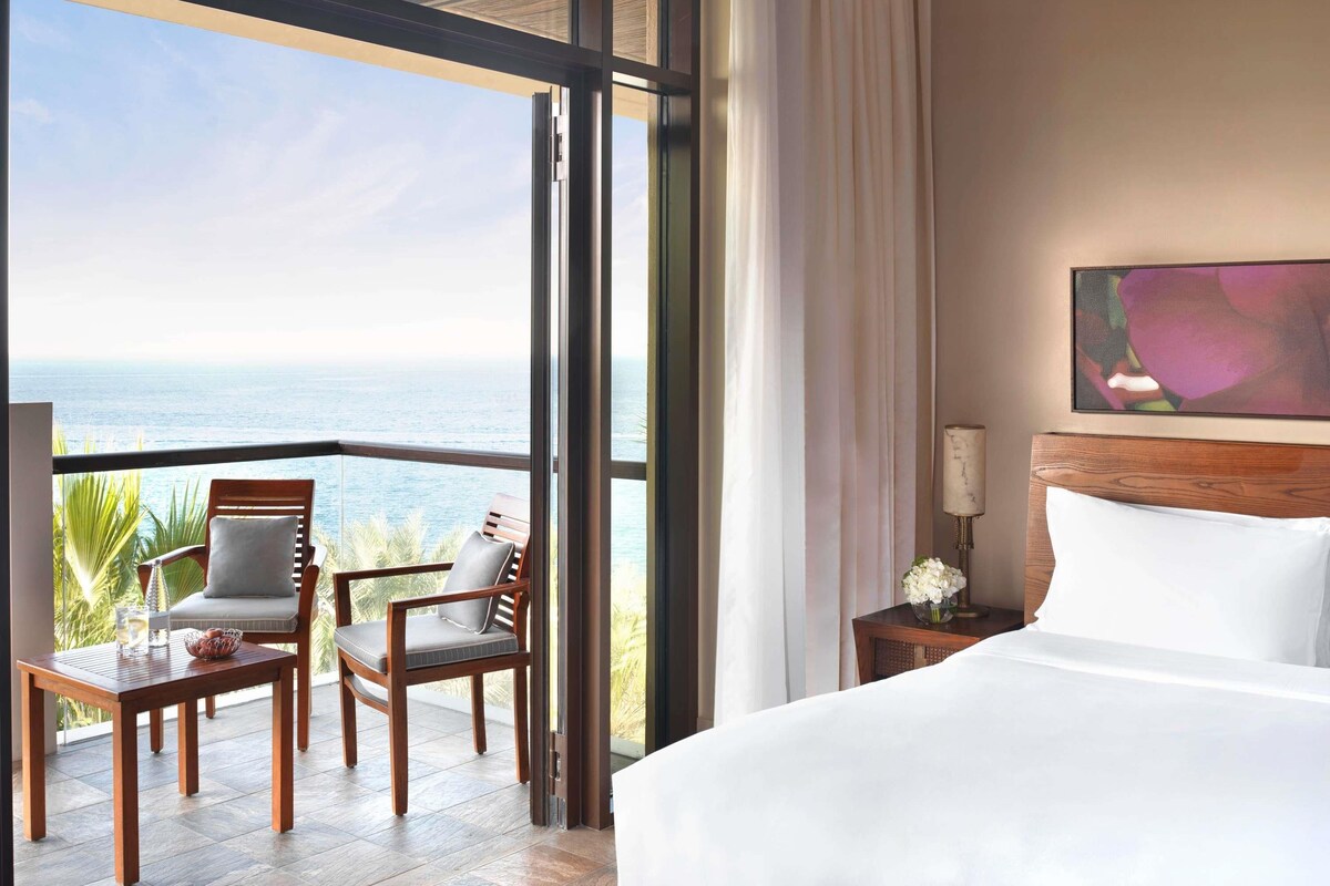 Een hotelkamer met een groot bed, een houten nachtkastje, een tafel met stoelen en uitzicht op zee.