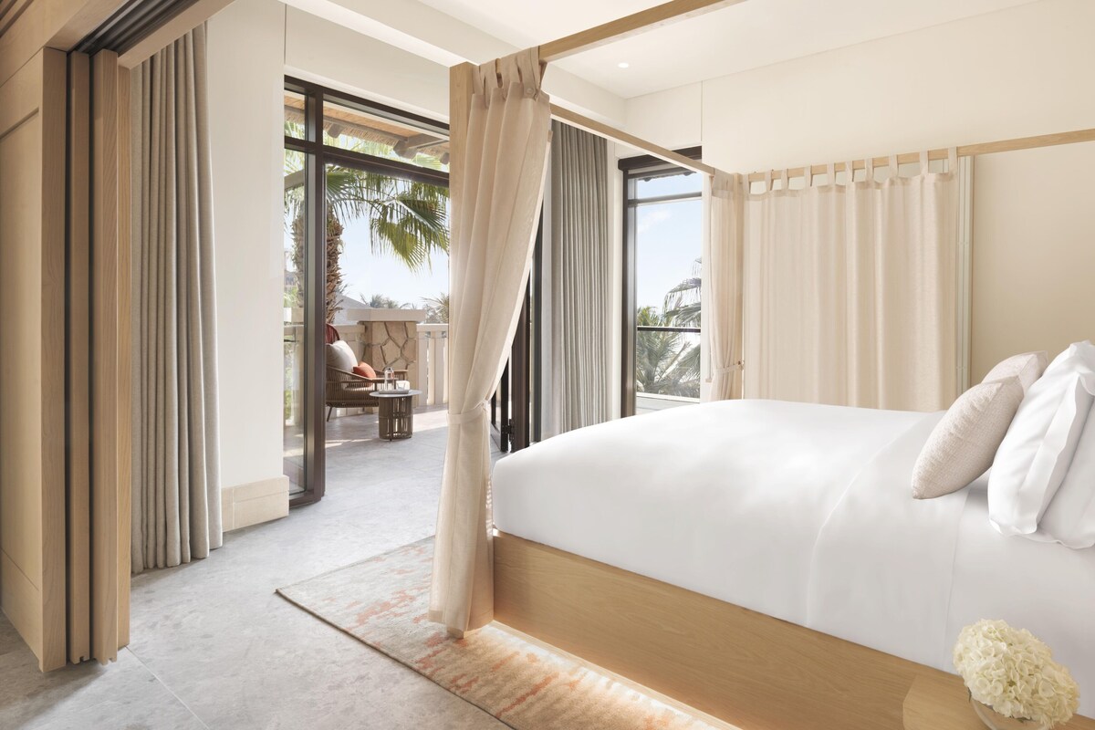 A modern bedroom with a large bed, a balcony with a view, and a seating area with a table and chairs.