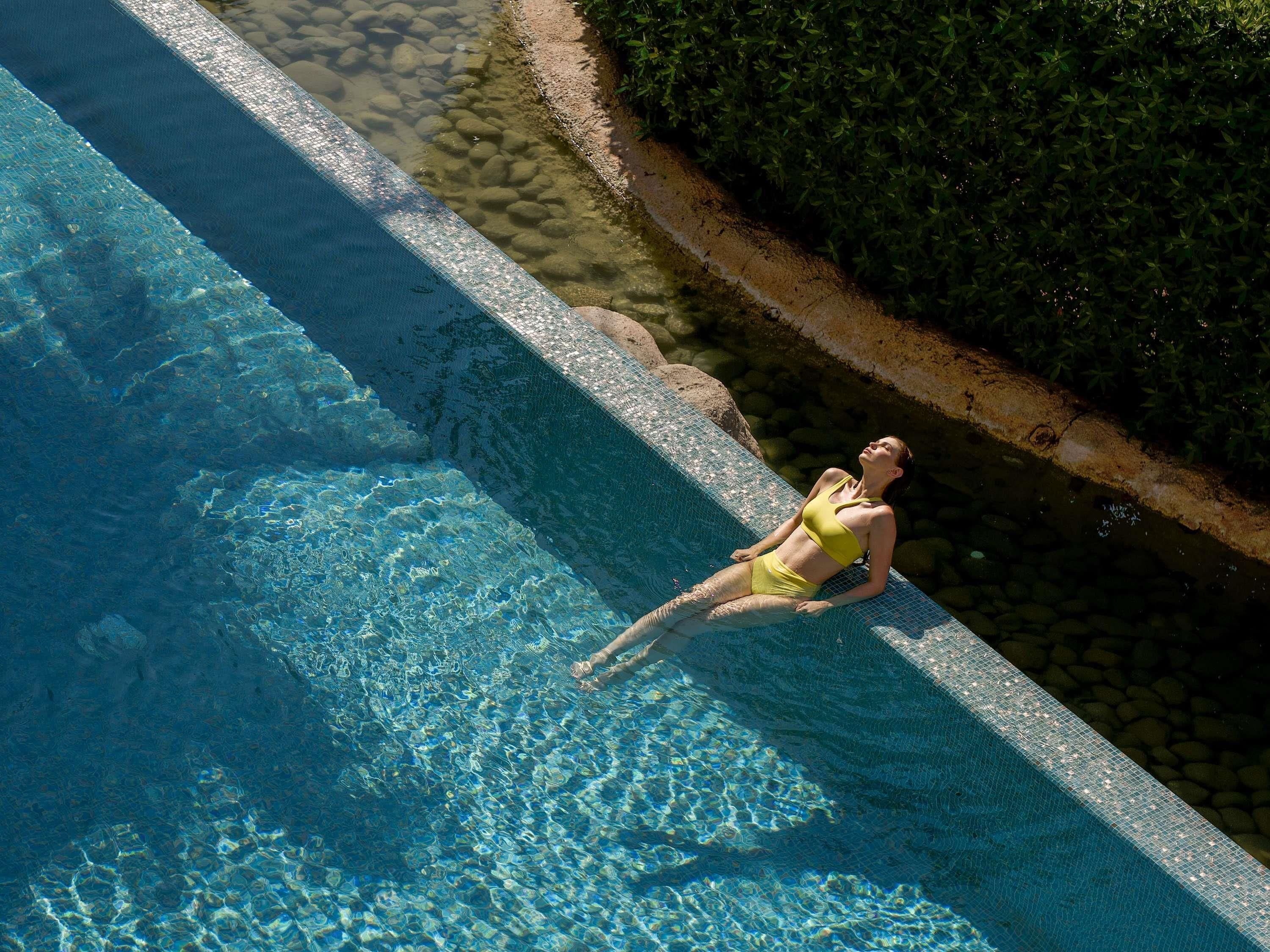Une femme vêtue d’un maillot de bain jaune se détend au bord d’une piscine à l’eau d’un bleu limpide, avec une rive rocheuse et une végétation luxuriante en arri ère-plan.