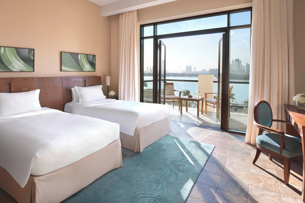 A hotel room with two beds, a balcony with a view of the city, and a table with chairs.