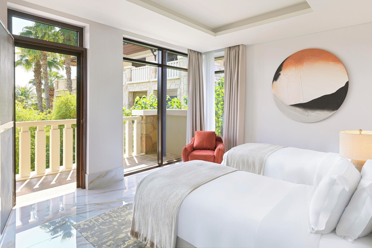 A hotel room with a large sliding glass door leading to a balcony, a round mirror on the wall, and a bed with white linens.