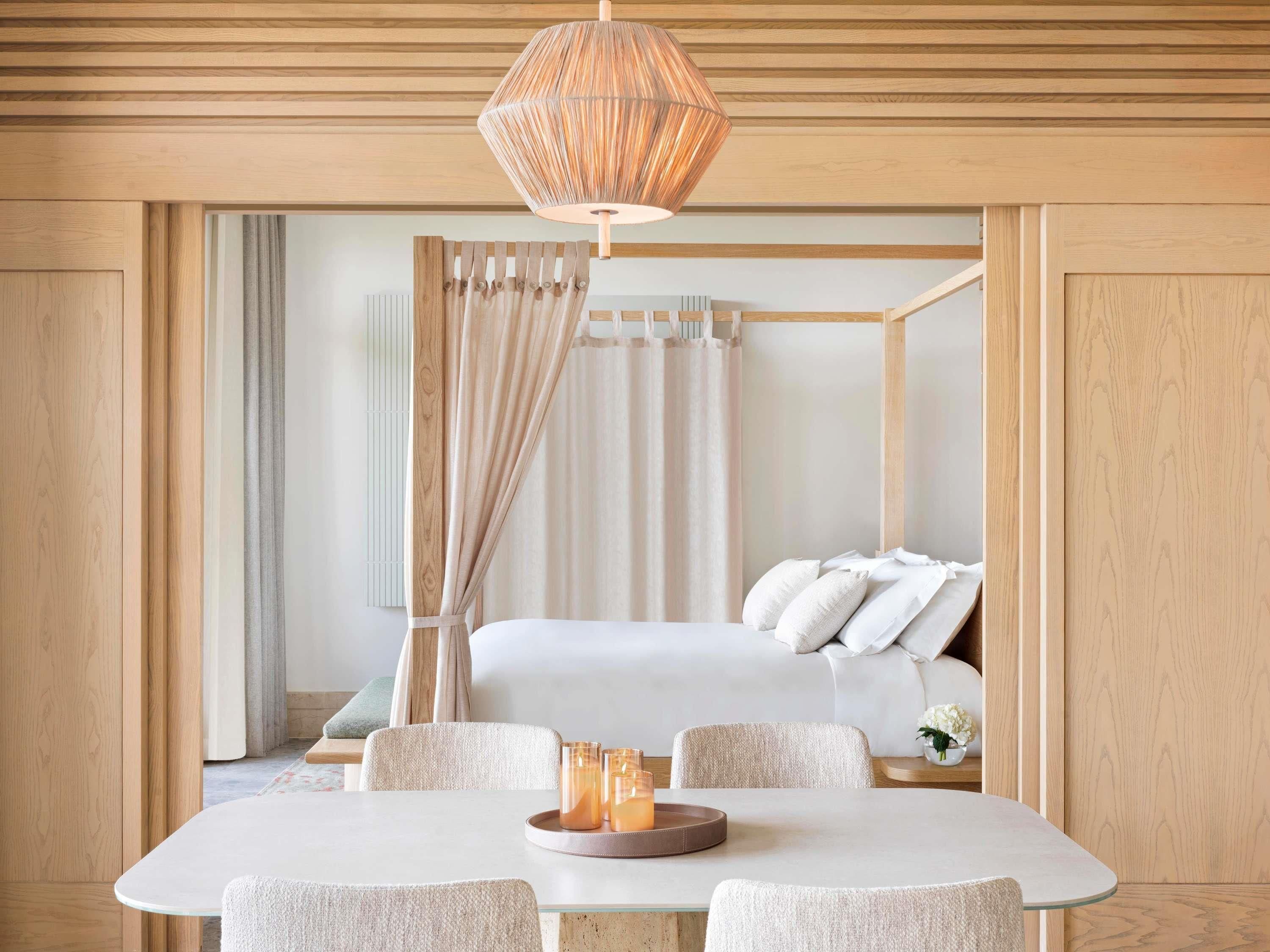 A modern bedroom with a wooden four-poster bed, a dining table with chairs, and a hanging light fixture.