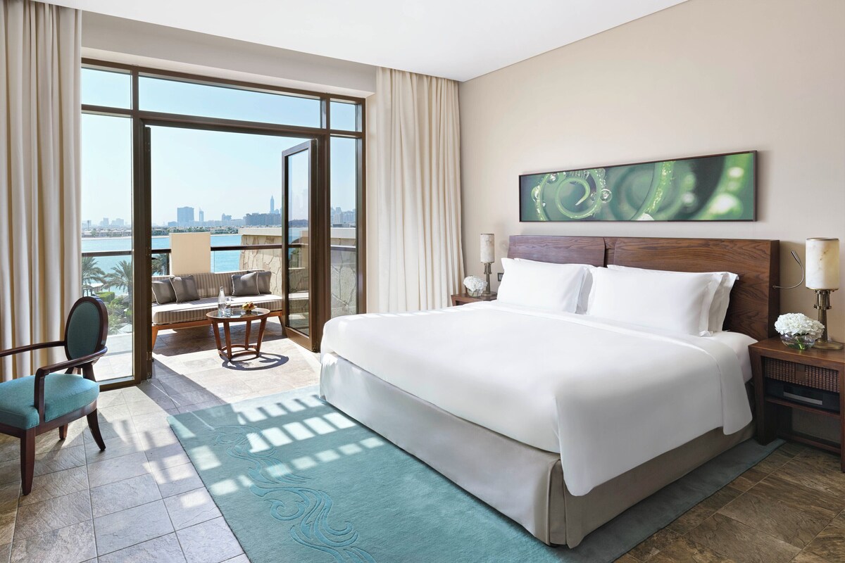 A hotel room with a large bed, a desk, a chair, a balcony with a view of the city, and a painting on the wall.