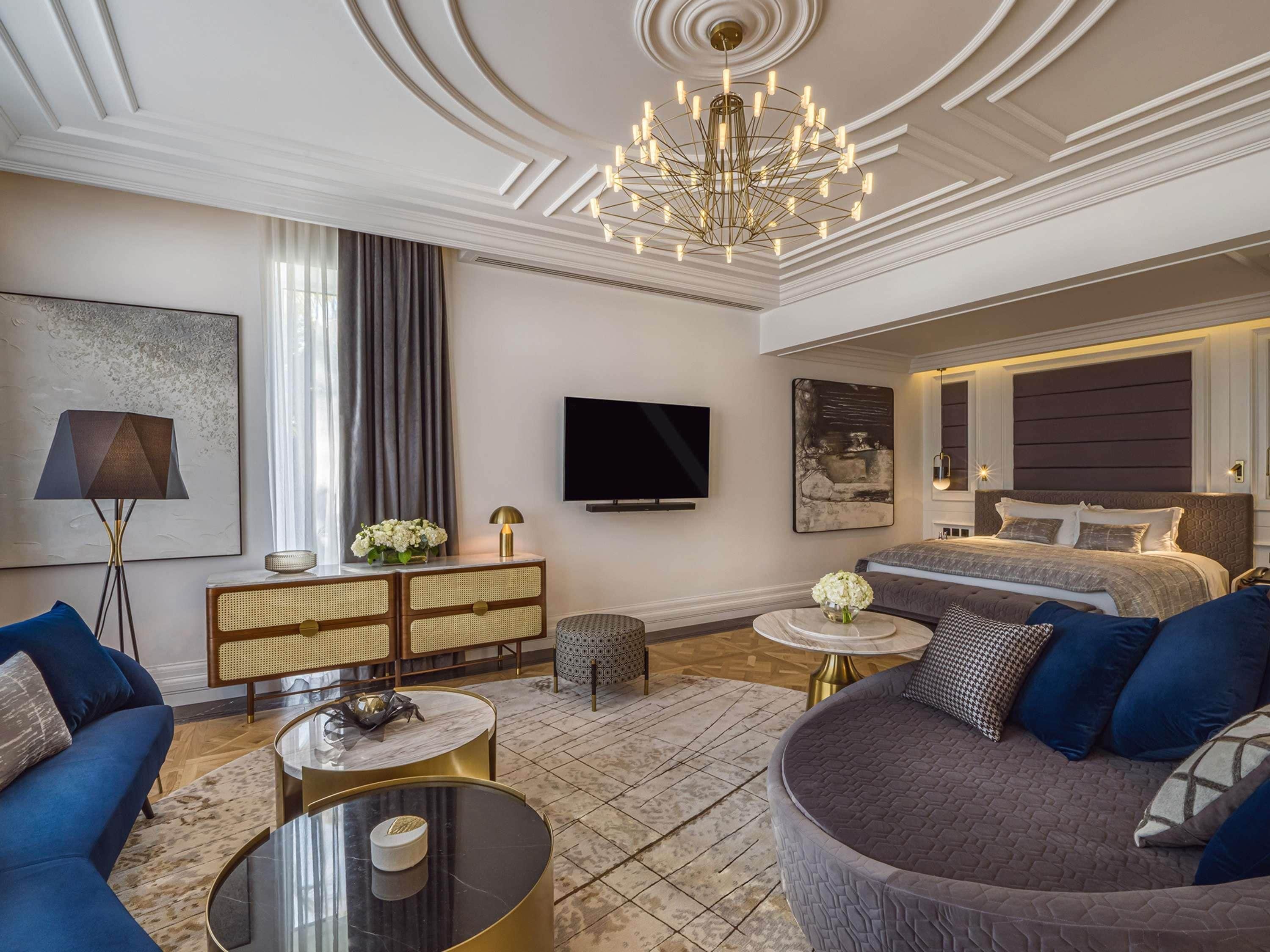 A modern hotel room with a large bed, a sofa, a TV, and a chandelier.