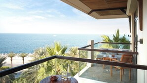 A balcony with a view of the sea, a table with chairs, and a small garden area with palm trees.