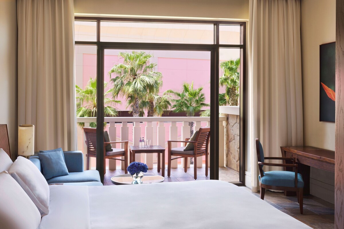 A hotel room with a large bed, a desk, and a balcony with a view of palm trees and a pink building.