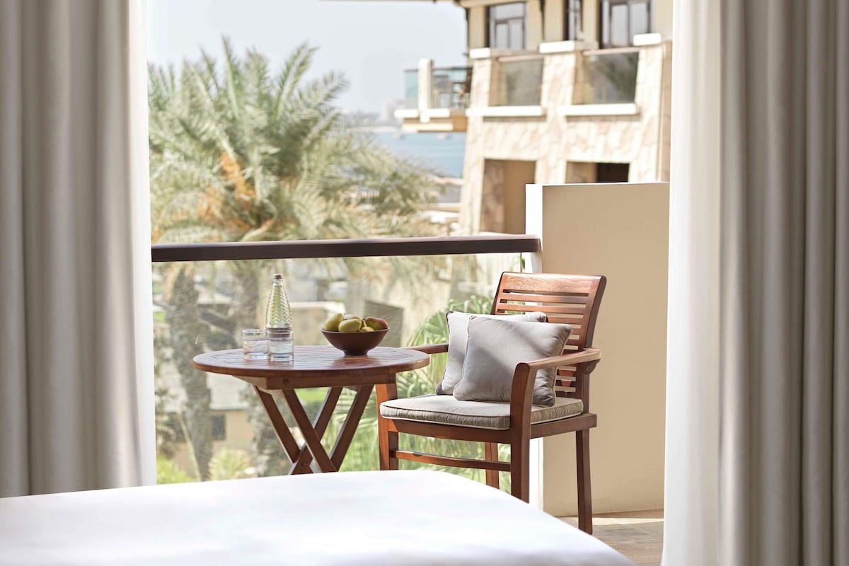 A balcony with a wooden chair, a small round table with a bowl and glasses, and a view of palm trees and a building.