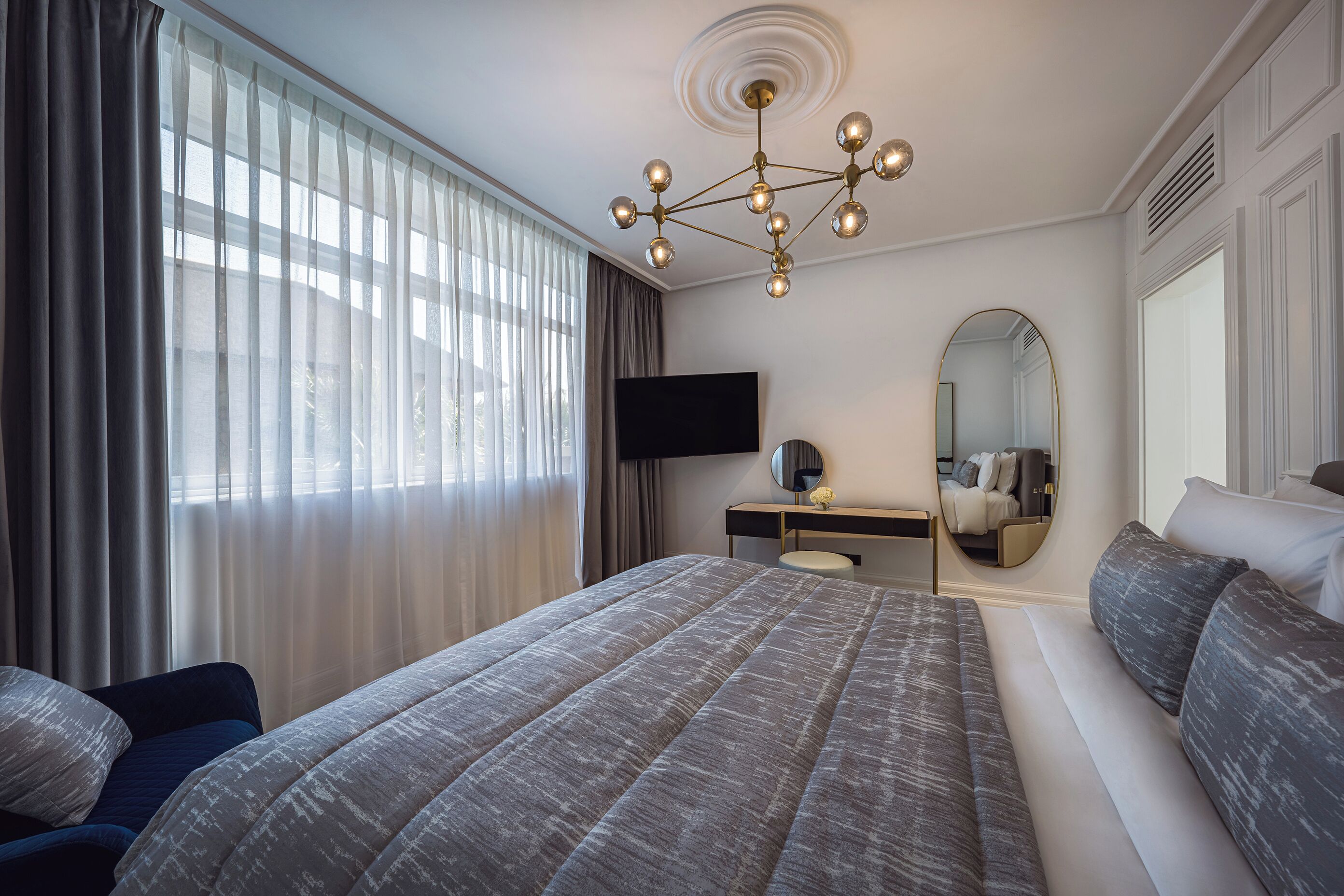 A modern hotel room with a large bed, a desk with a flat-screen TV, a mirror, and a chandelier.