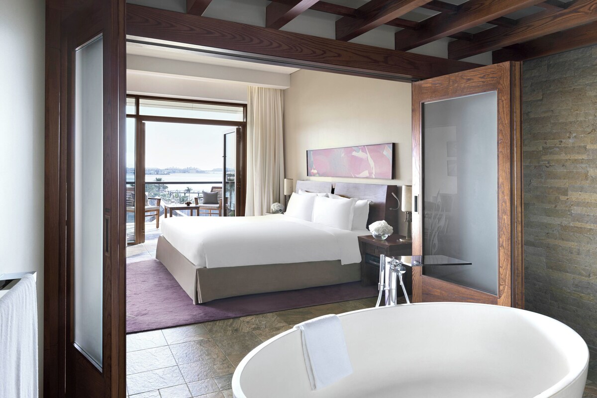 Una habitación de hotel moderna con una cama grande, un baño con bañera independiente y un balcón con vistas a la ciudad.