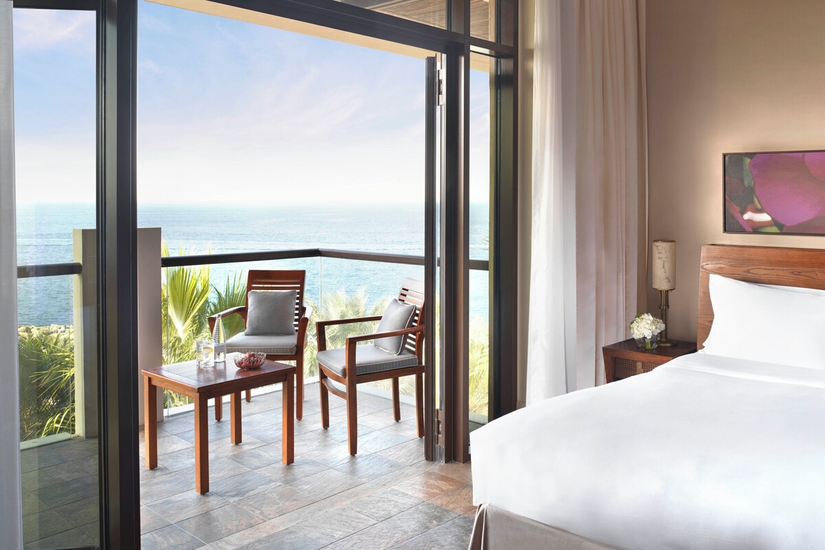 A hotel room with a large bed, a view of the ocean, a dining area with chairs, and a painting on the wall.