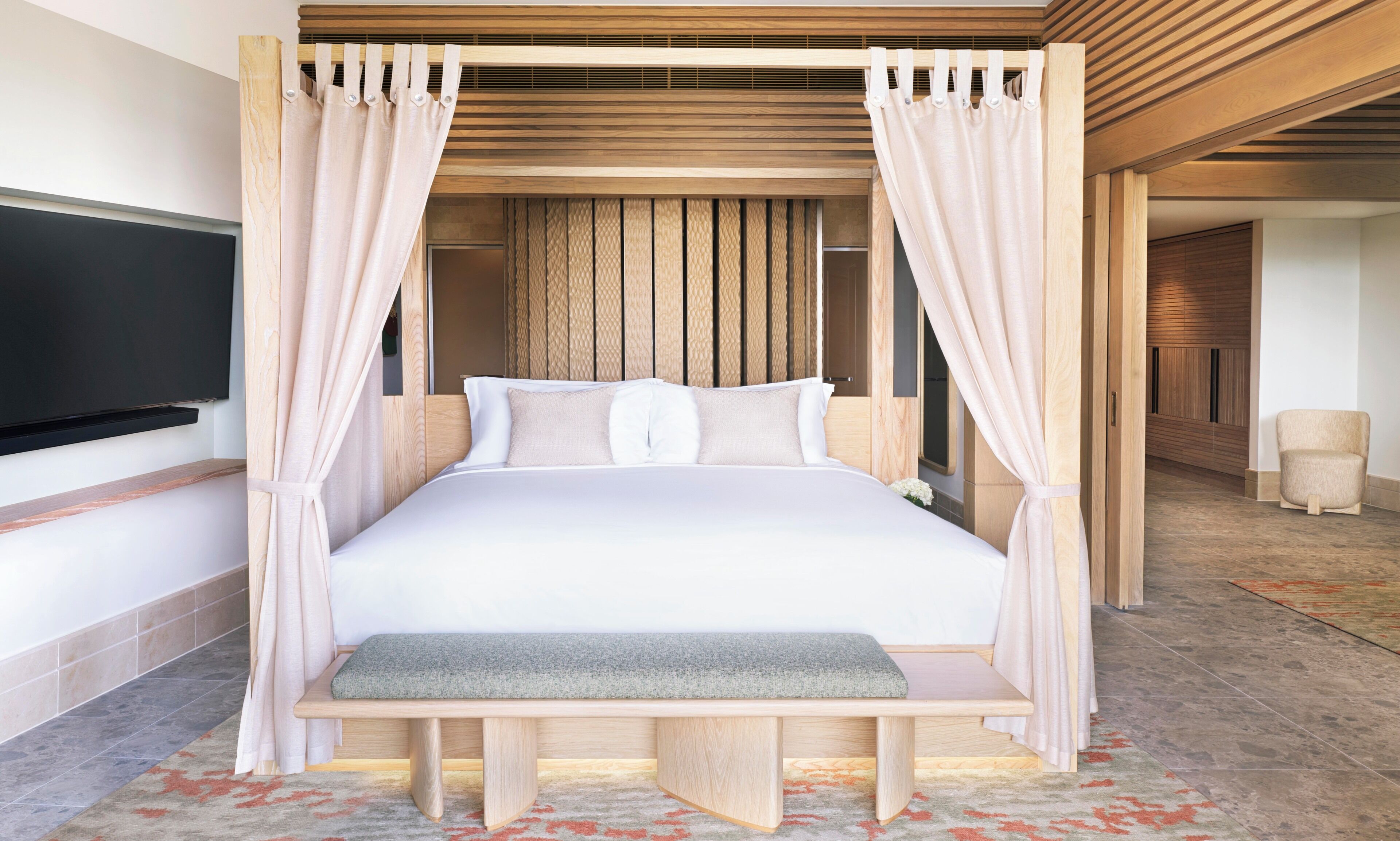 A bedroom with a canopy bed, a bench, a TV, and a bathroom visible in the background.