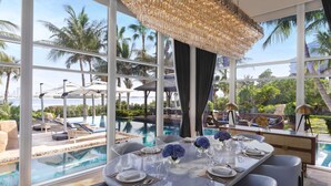 Un área de piscina interior lujosa con un gran candelabro, una mesa de comedor para cuatro personas y vistas al océano y palmeras.