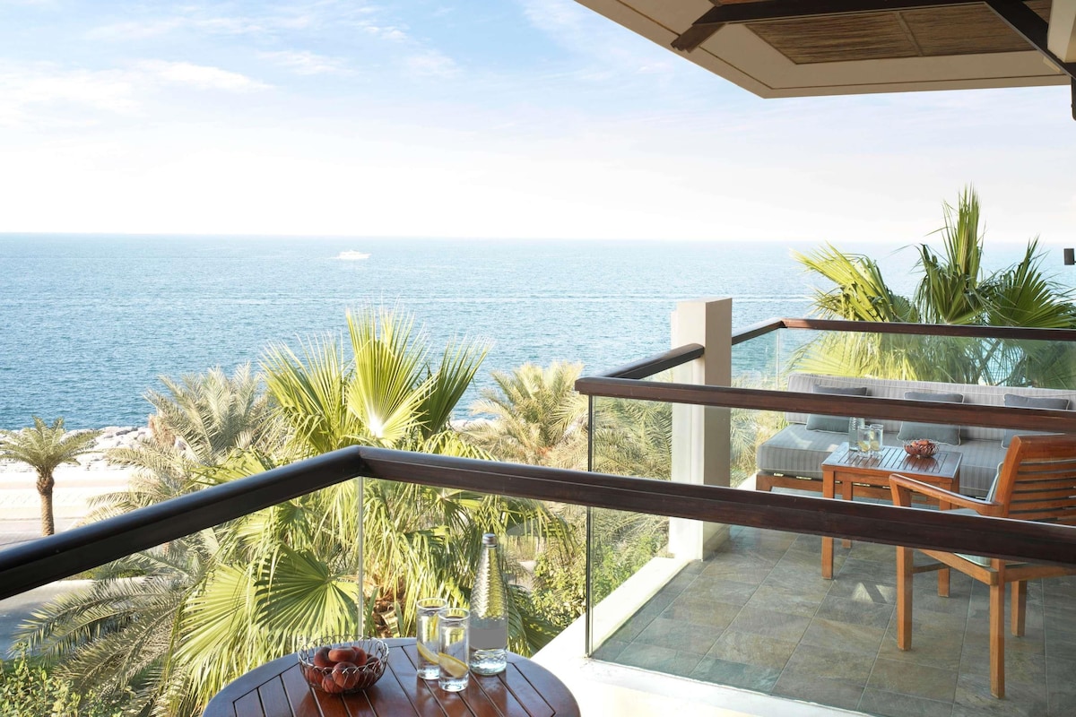 Un balcón con vistas al mar, amueblado con una mesa y sillas, y barandilla de vidrio.