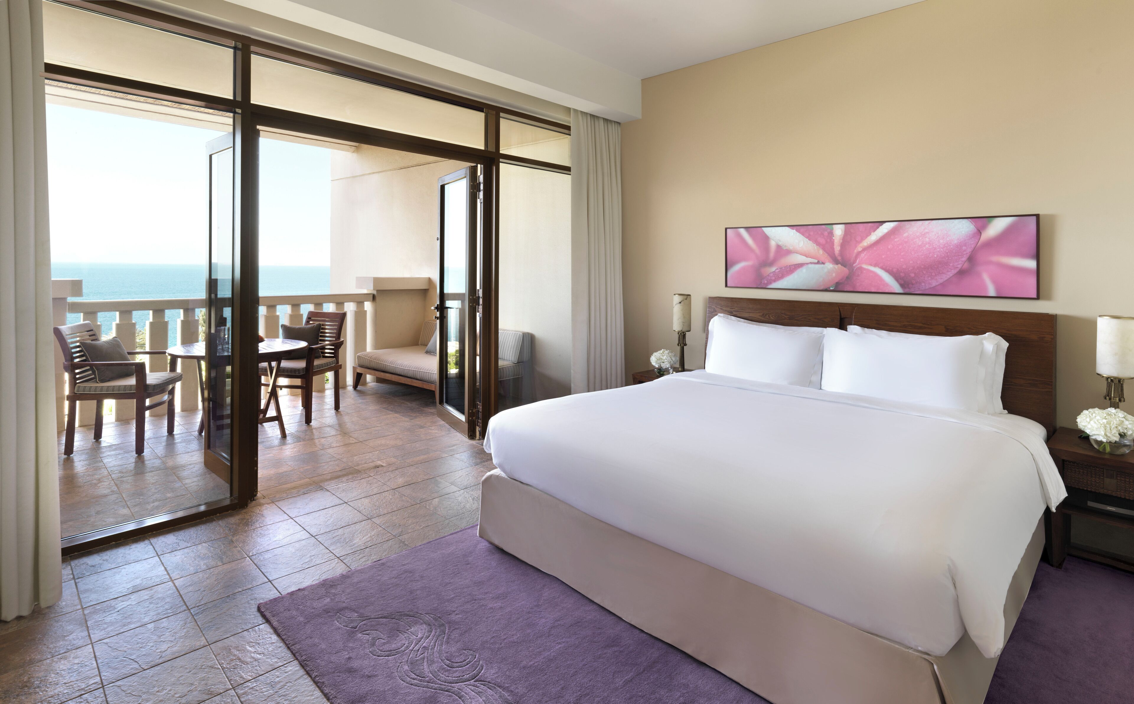 A hotel room with a large bed, a balcony with a view of the sea, a dining area with chairs, and a painting on the wall.