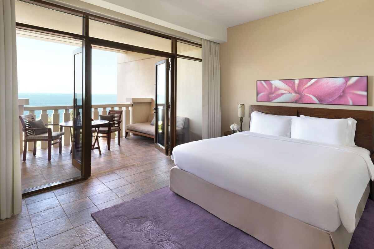 A hotel room with a large bed, a balcony with a view of the sea, a dining area with chairs, and a painting on the wall.