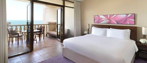 A hotel room with a large bed, a balcony with a view of the sea, a dining area with chairs, and a painting on the wall.