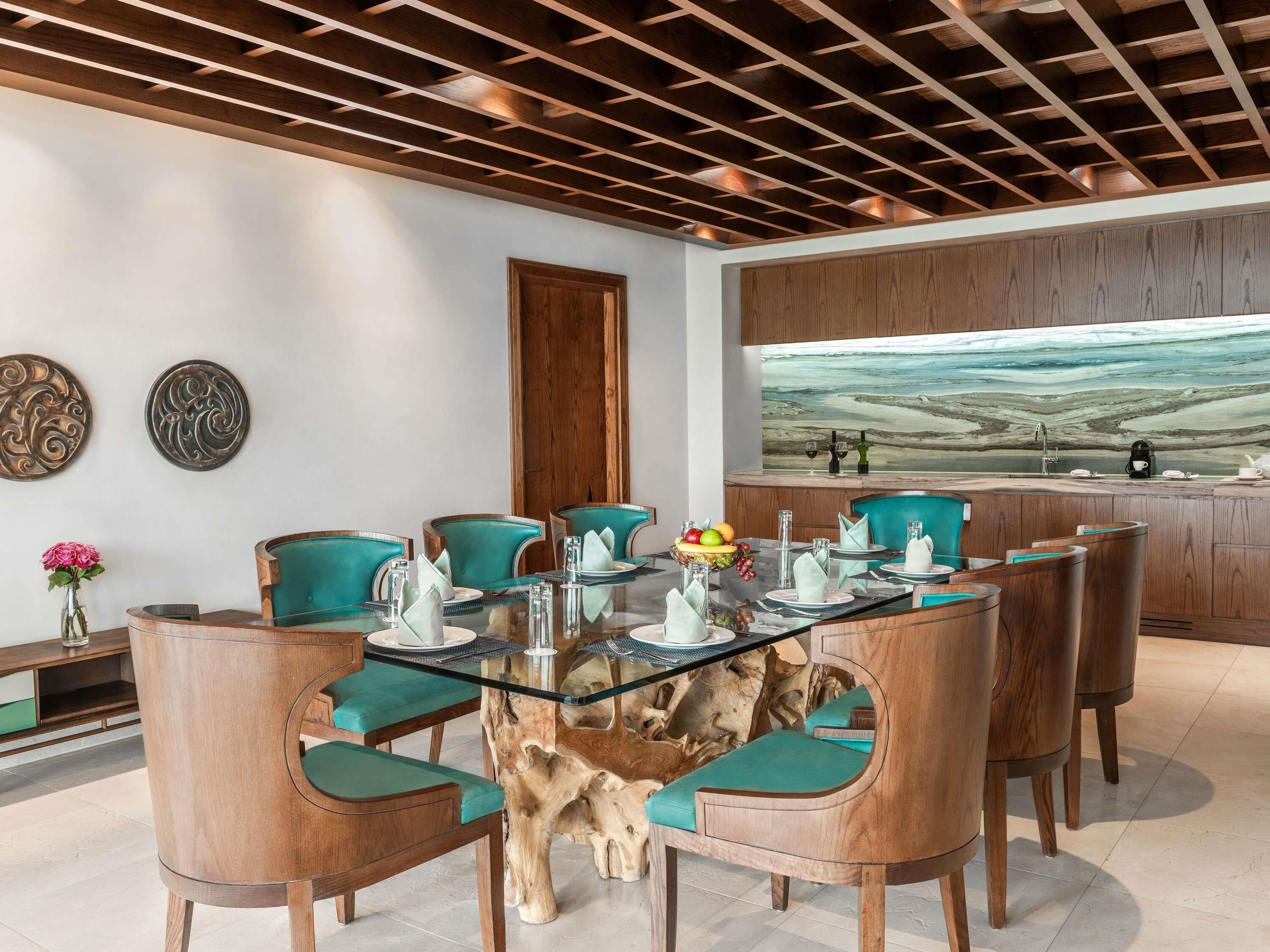 Un coin repas avec une table en verre, des chaises en bois dotées de coussins turquoise, un buffet en bois et un grand tableau abstrait accroché au mur.
