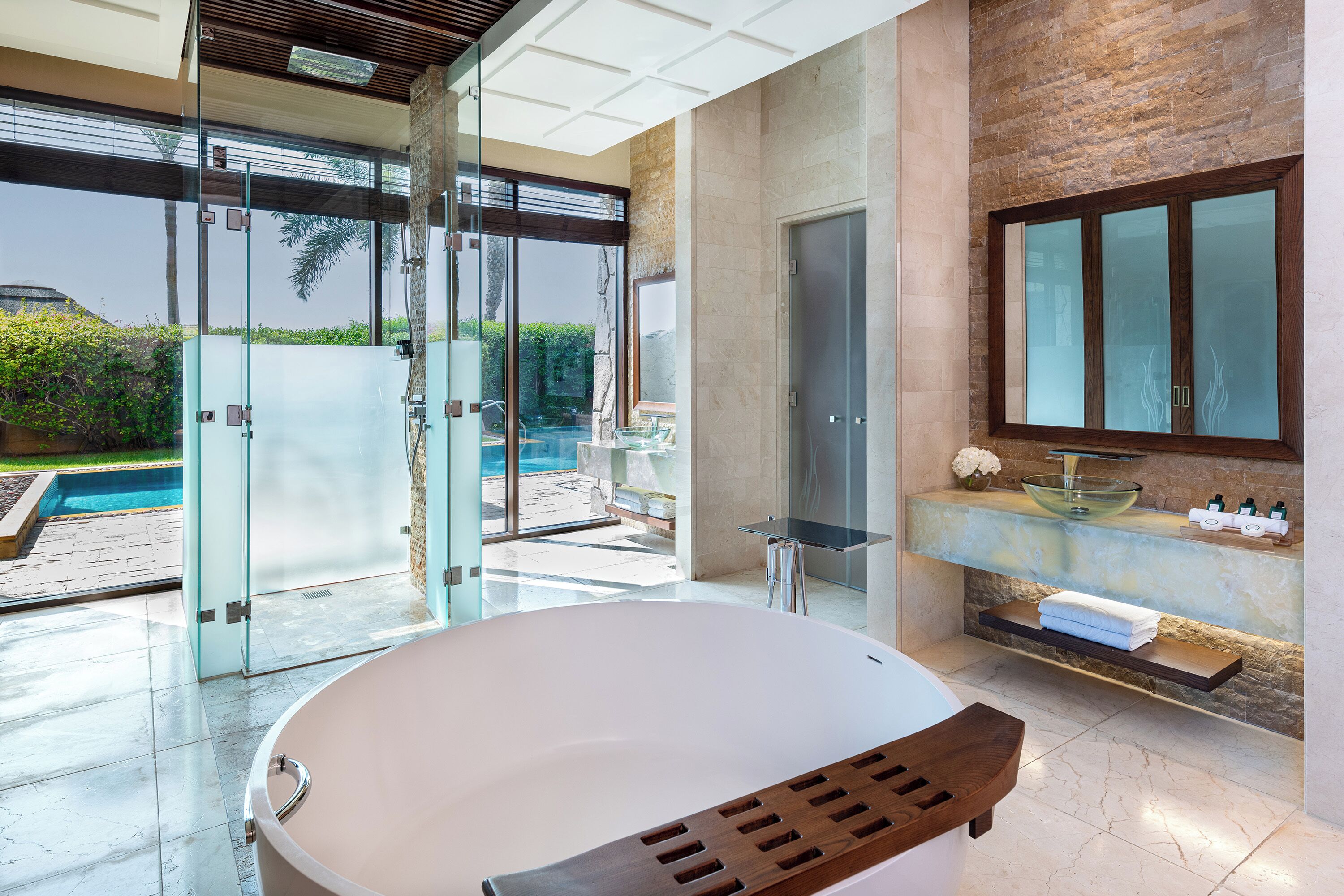 Une salle de bain moderne dotée d’une grande baignoire, de parois de douche en verre, d’un comptoir en marbre et offrant une vue sur une piscine.