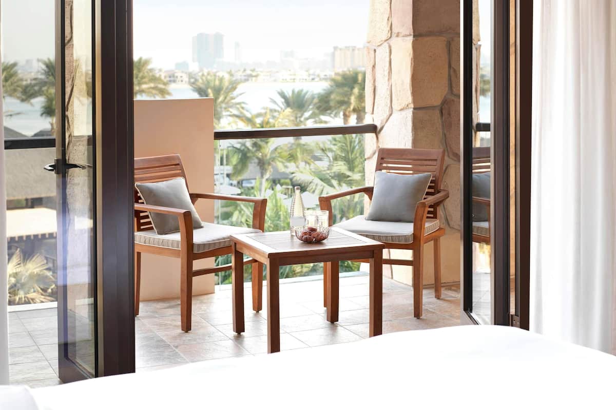 Een balkon met twee houten stoelen, een kleintje tafeltje en uitzicht op palmbomen en gebouwen.