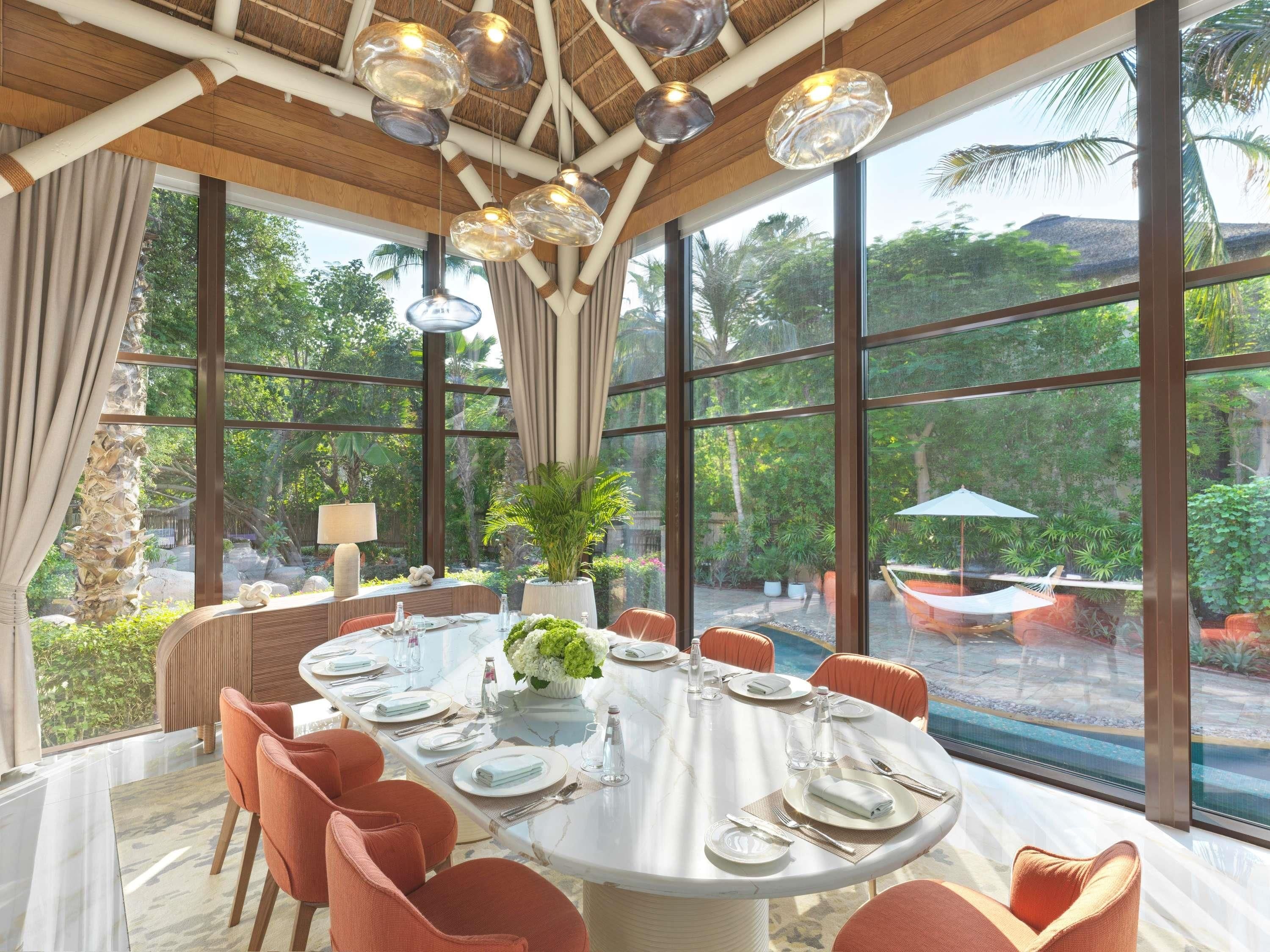 Un coin repas avec une table ronde dressée pour six personnes, entourée de chaises orange, de grandes fenêtres et offrant une vue sur un jardin tropical.