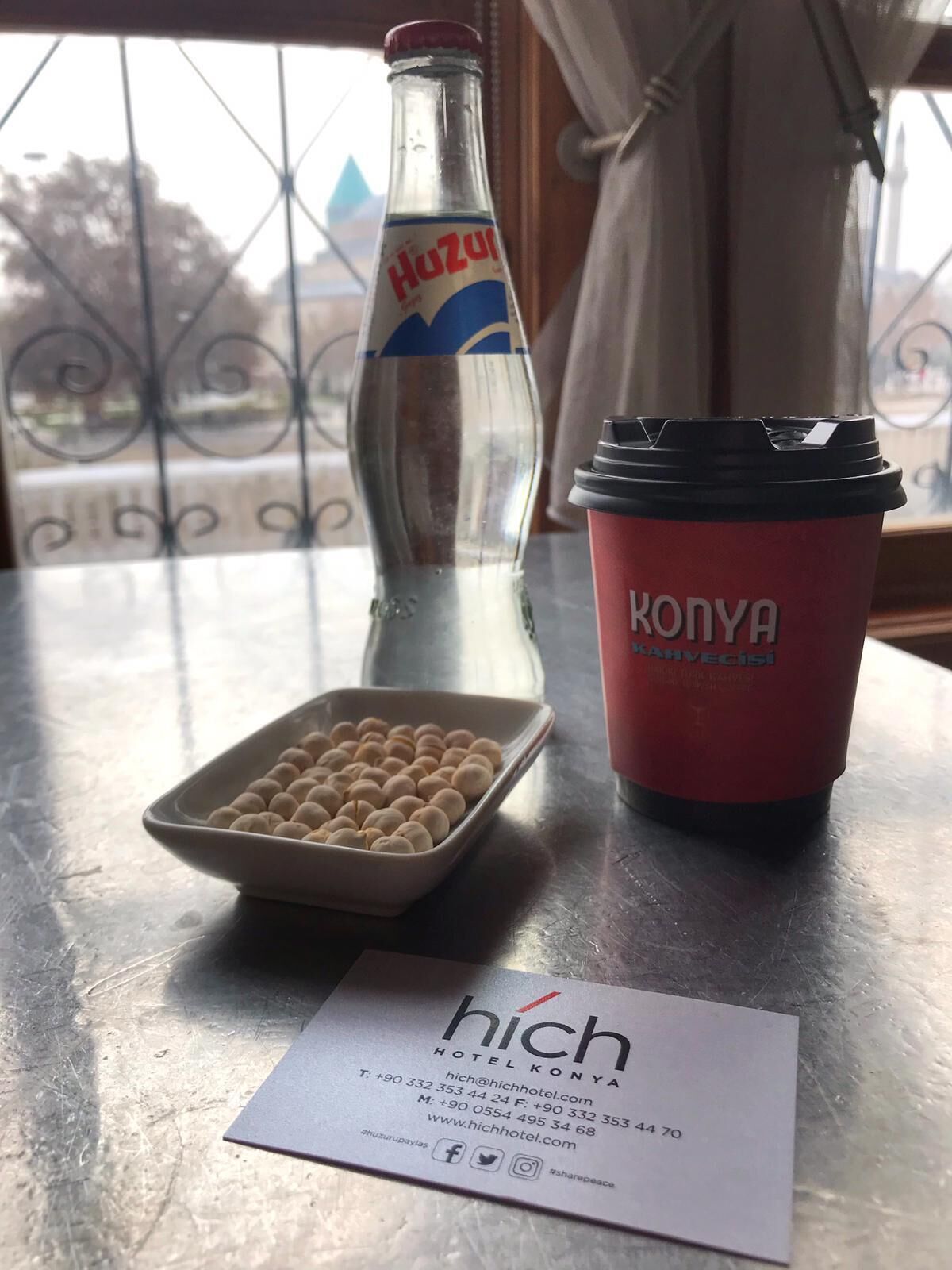 Eine Flasche Huzural, eine rote Tasse mit dem Schriftzug „KONYA“ sowie eine Schale mit runden Gegenständen und eine Visitenkarte für das „HICH HOTEL KONYA“ mit Kontaktdaten.