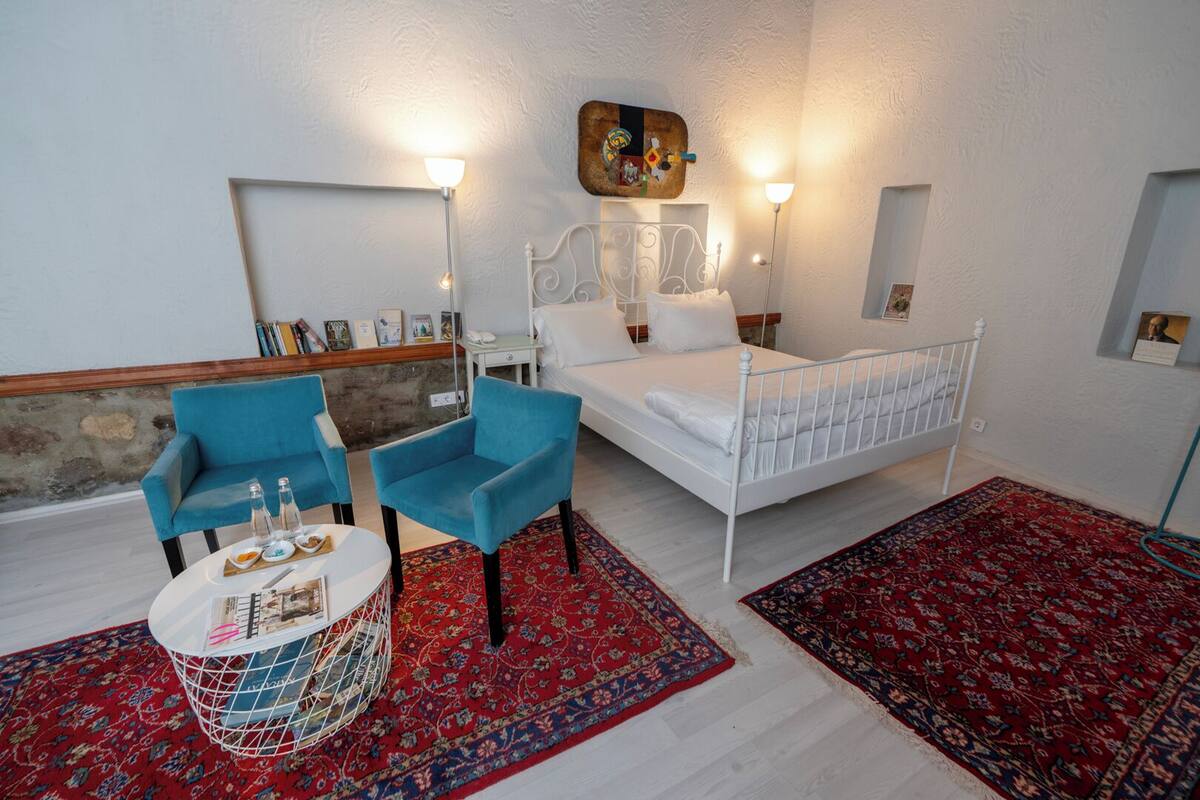 A bedroom with a white bed, two blue armchairs, a small round table with a tray, and two red patterned rugs.