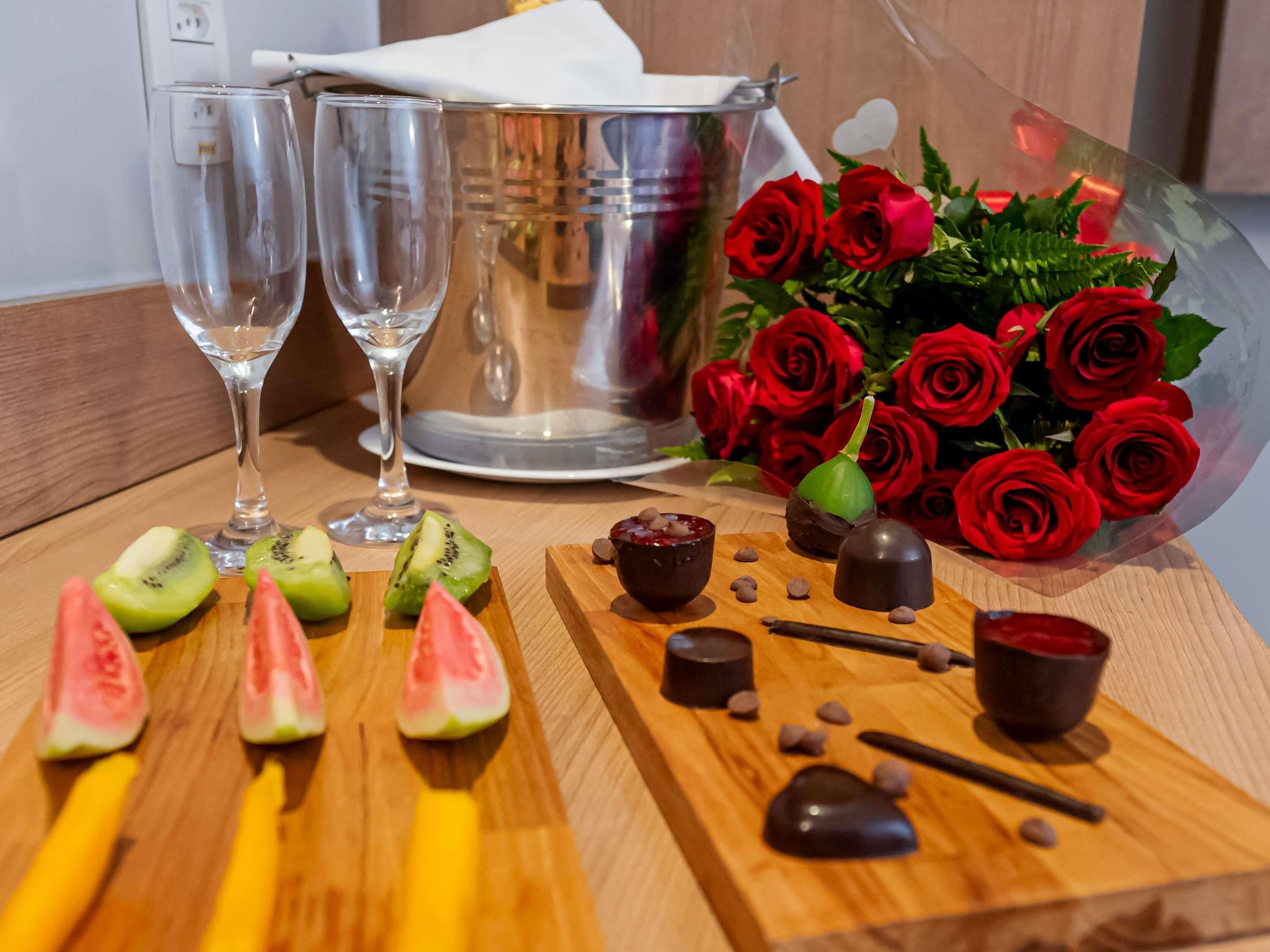 A wooden table with sliced fruits, chocolate treats, and a bouquet of red roses.
