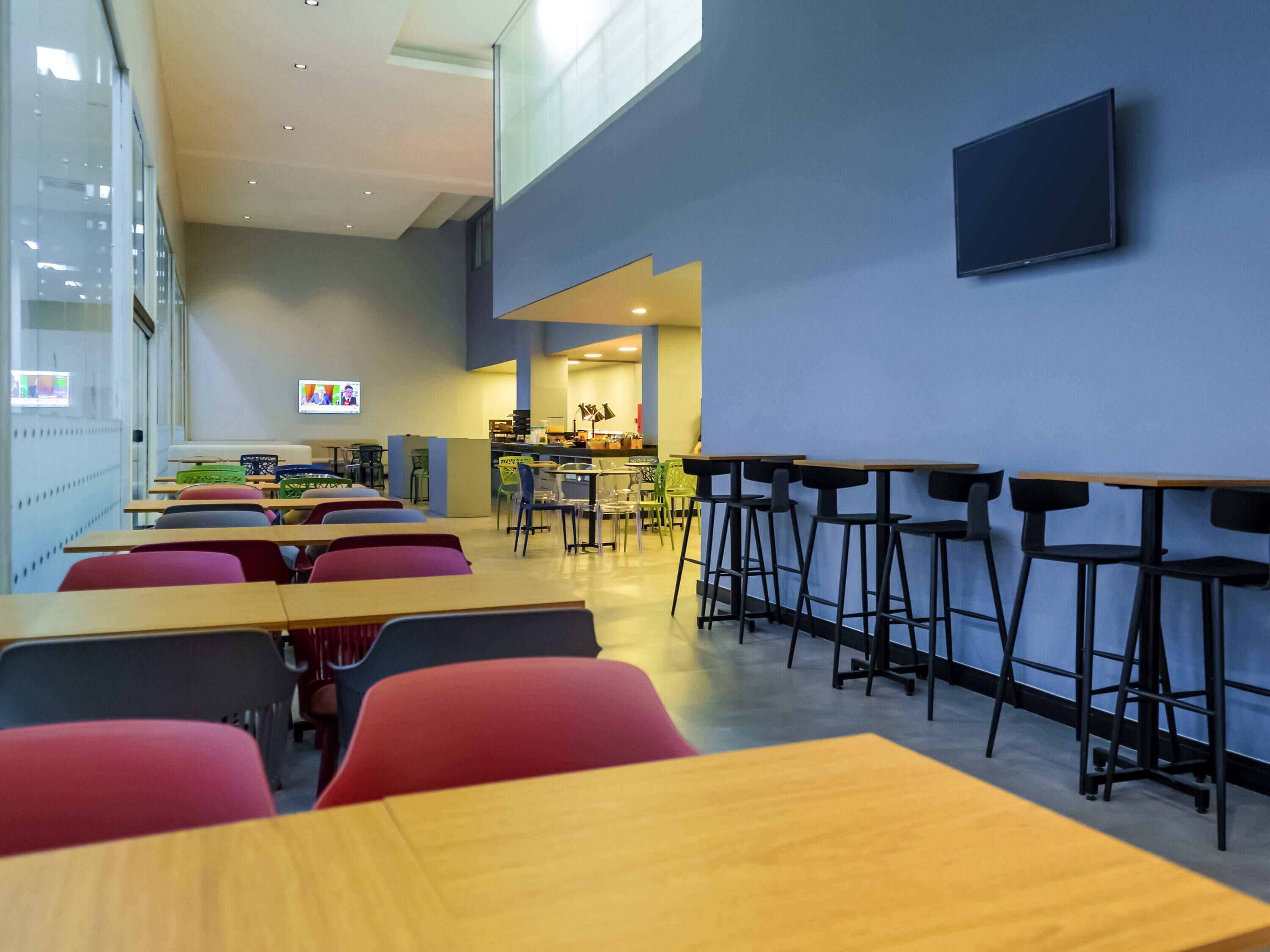 A modern dining area with high tables and chairs, a kitchen in the background, and a TV mounted on the wall.
