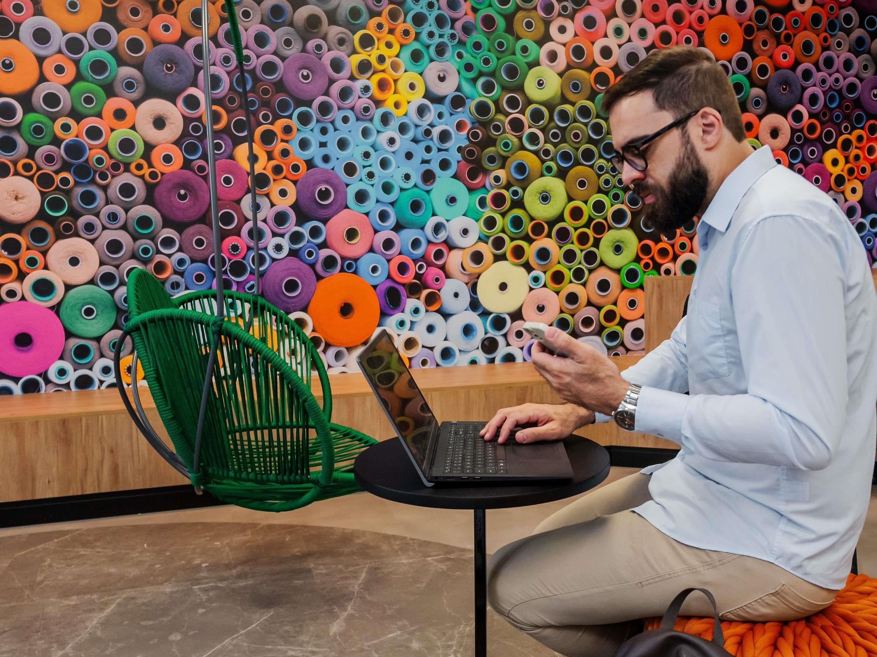 Una persona sentada en una silla frente a una mesa con una computadora portátil, frente a un muro lleno de ovillos de colores.