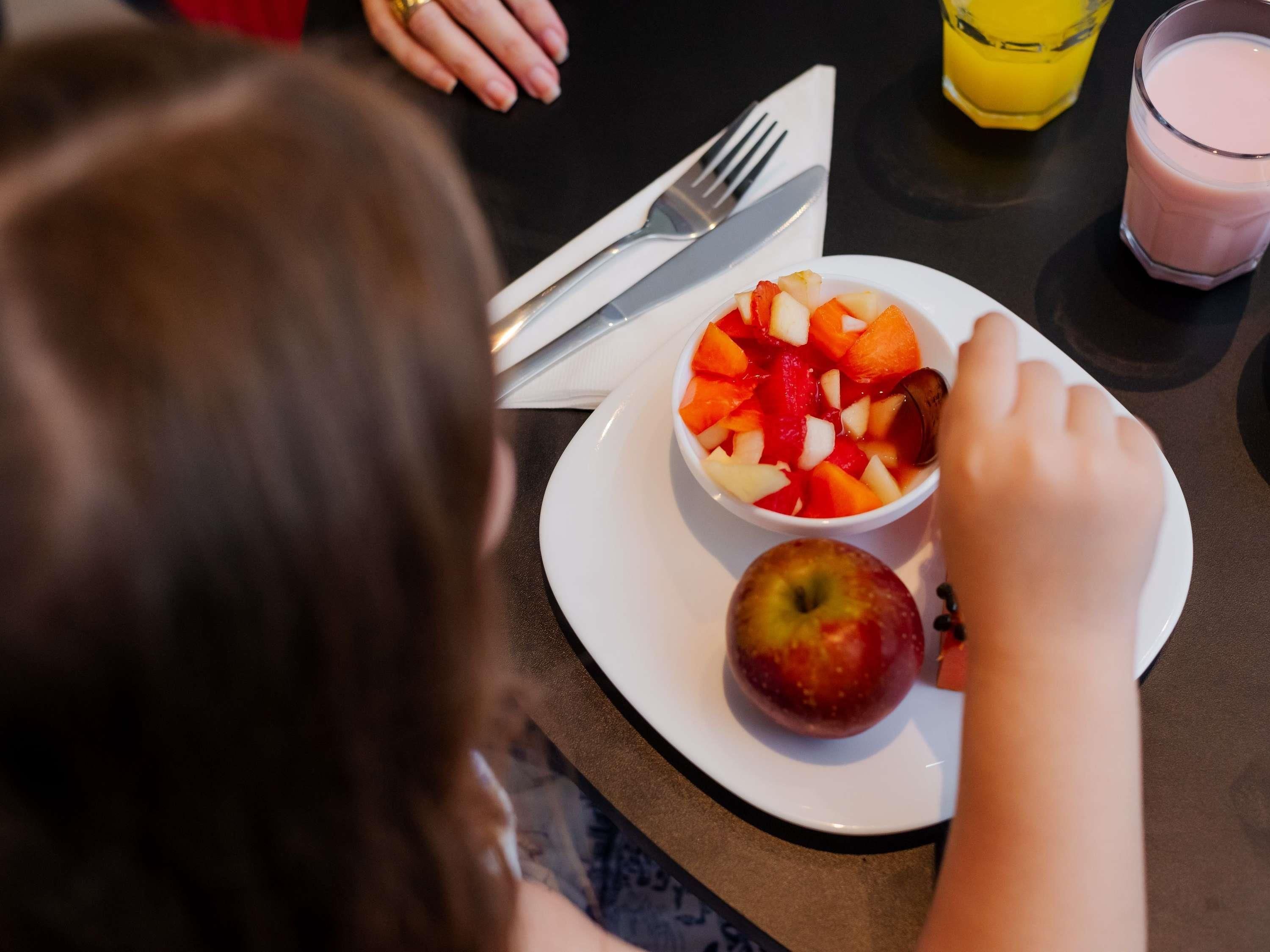 A dining table with a white plate containing a fruit salad and an apple, a fork, a knife, and a glass of juice.