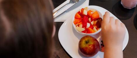 A dining table with a white plate containing a fruit salad and an apple, a fork, a knife, and a glass of juice.
