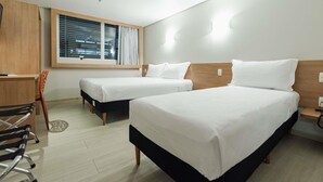 Une chambre d’hôtel avec deux lits, un bureau, une chaise, une fenêtre avec des stores et une vue sur le paysage urbain.