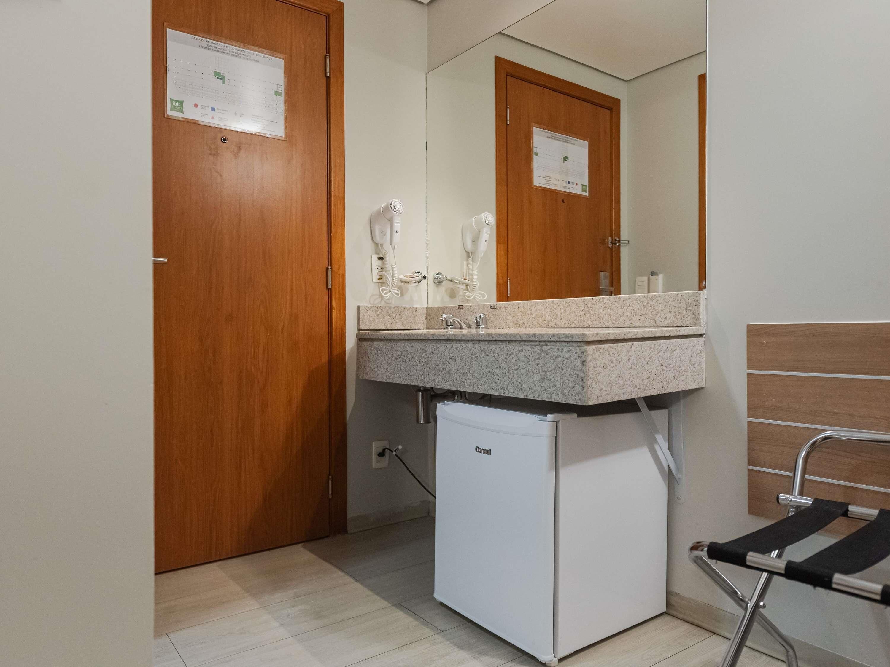 Un baño con lavamanos, espejo, secador de pelo y un refrigerador pequeño.