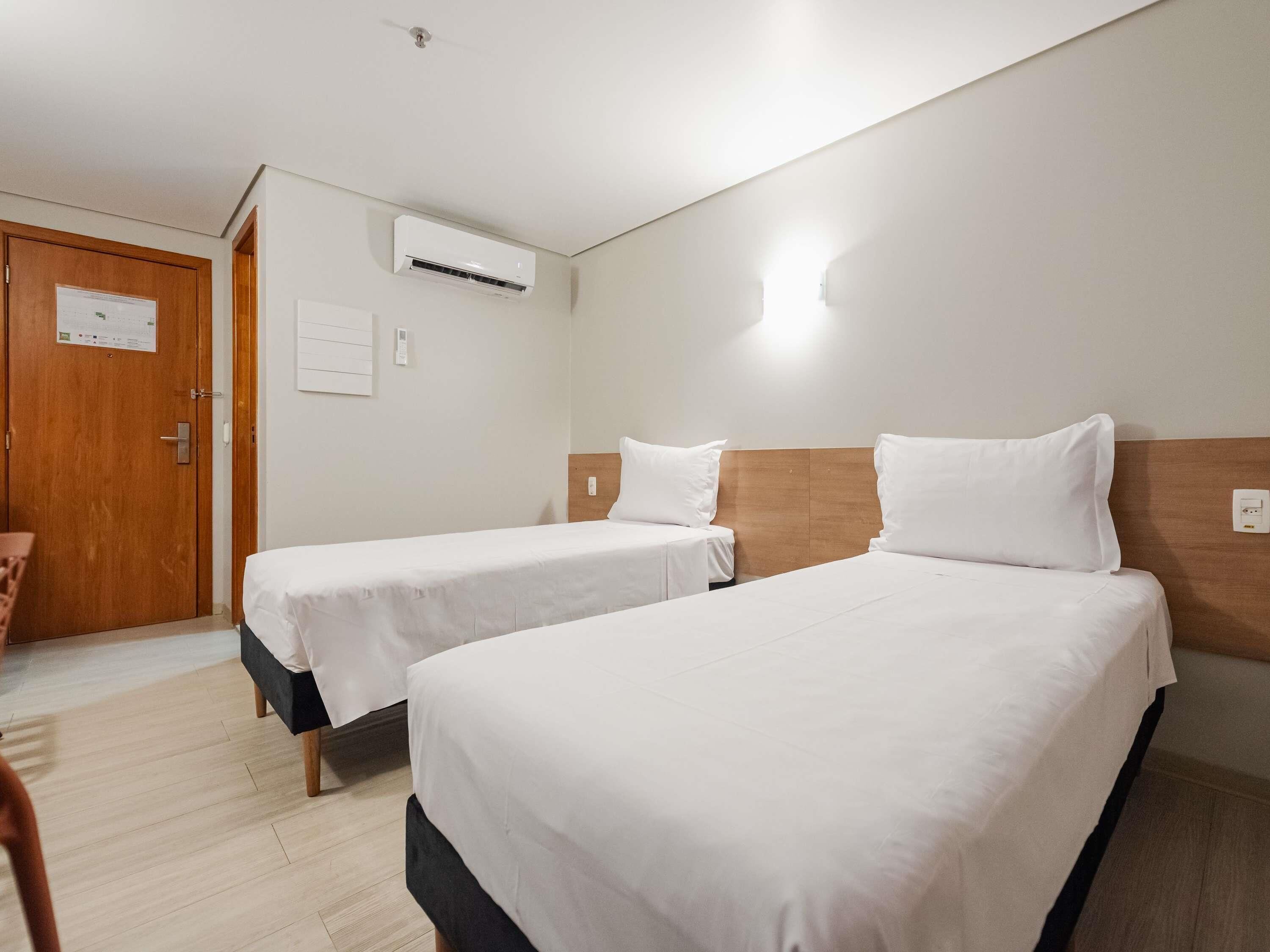 Habitación de hotel con dos camas individuales, una puerta de madera, un equipo de aire acondicionado y un interruptor de luz en la pared.