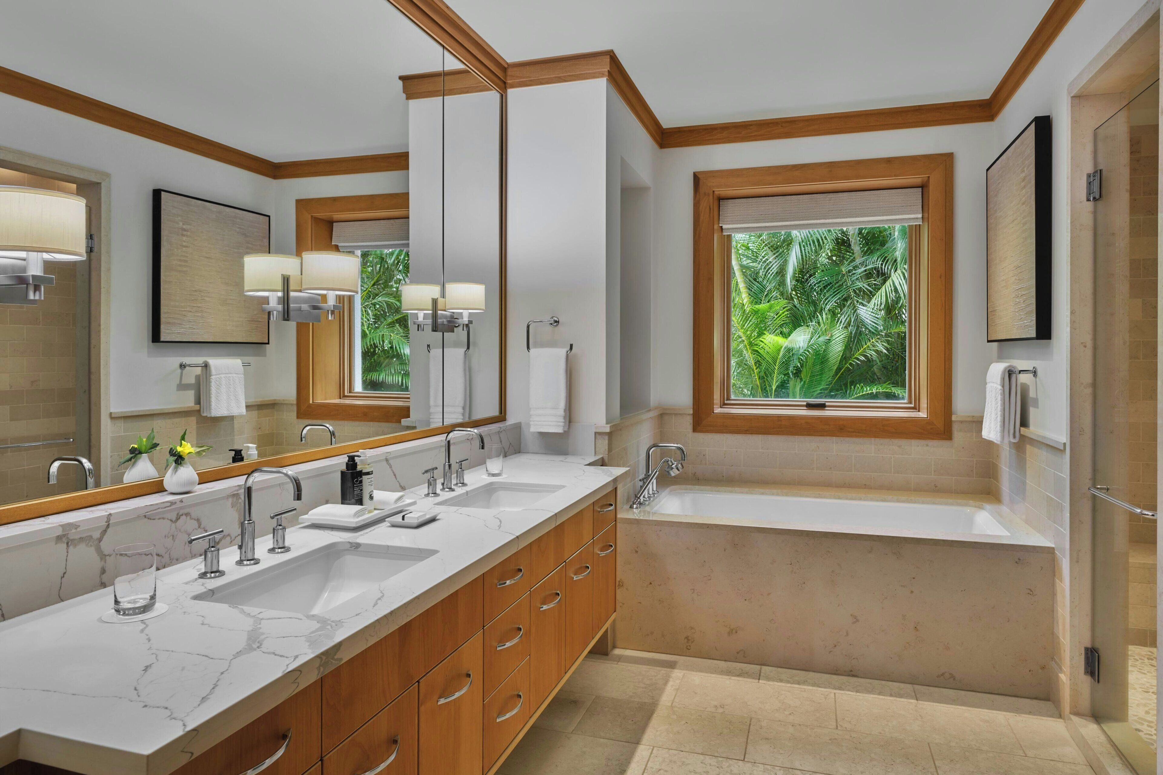 Un baño moderno con un espejo grande, dos lavamanos, una bañera y una ventana con vistas a la vegetación.