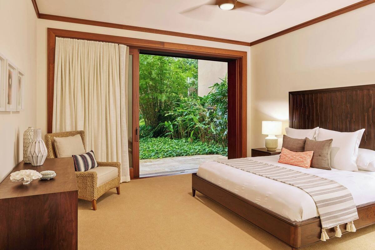 Een slaapkamer met een bed, een stoel, een bureau, een lamp en uitzicht op groen.
