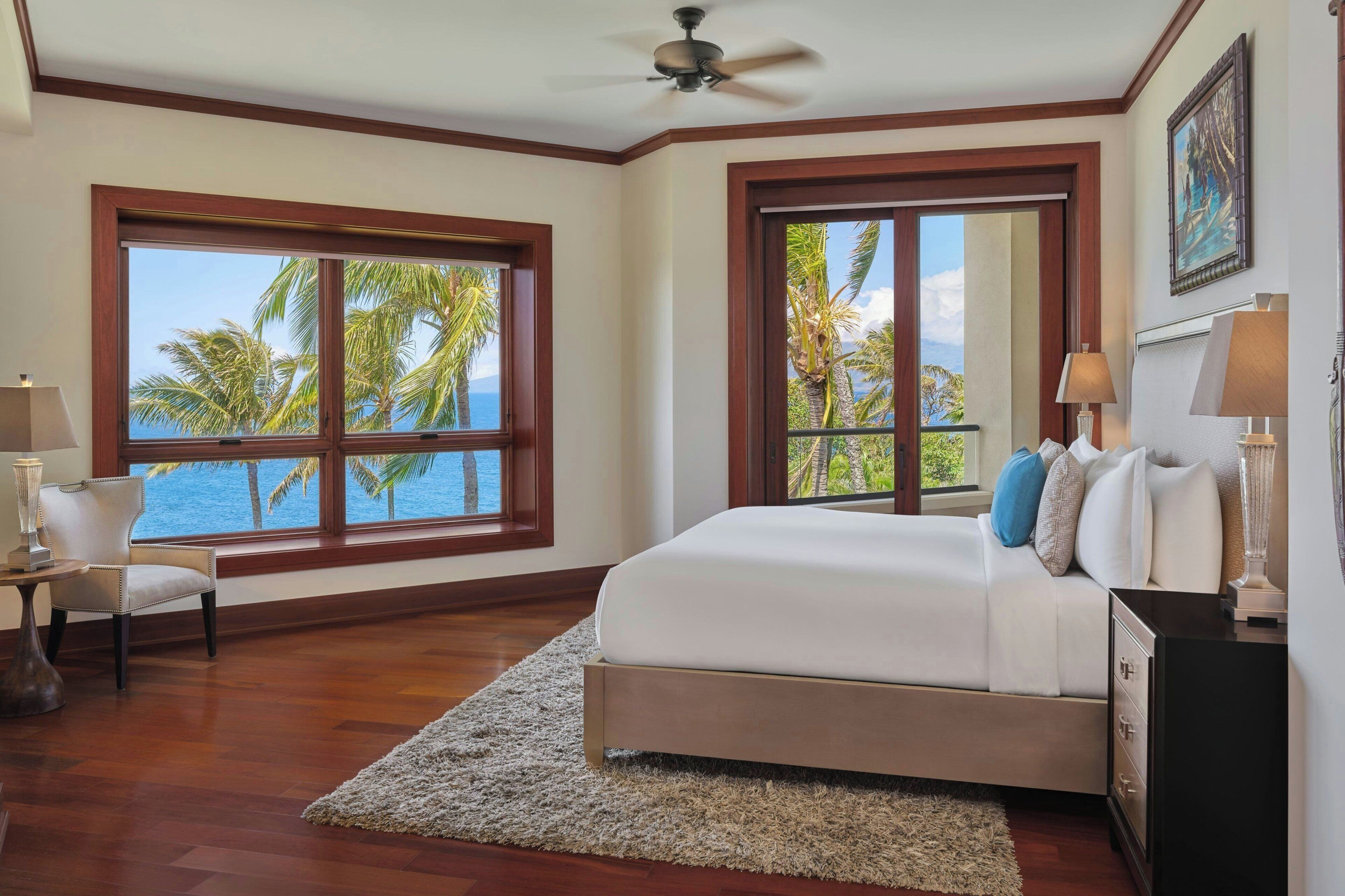 A bedroom with a large bed, two windows with ocean views, a ceiling fan, and a nightstand with a lamp.