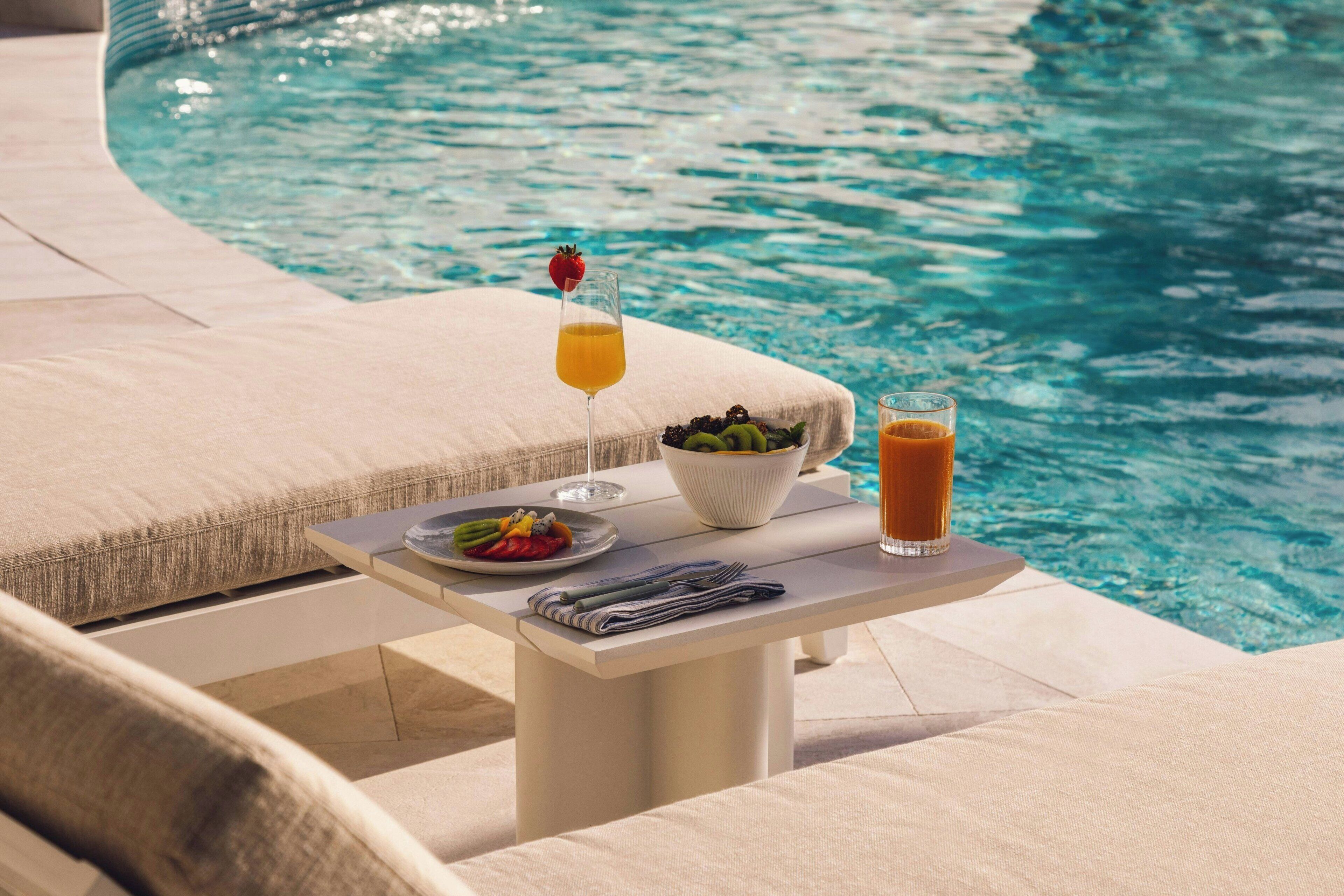Mesa junto a la piscina con un vaso de zumo de naranja, un bol de fruta y un vaso de té helado.