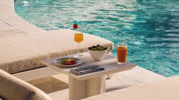 Mesa junto a la piscina con un vaso de zumo de naranja, un bol de fruta y un vaso de té helado.