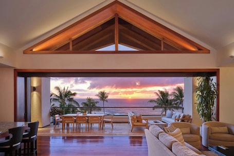 A spacious living area with a high ceiling, a dining set, and a sofa facing a large window overlooking the ocean at sunset.