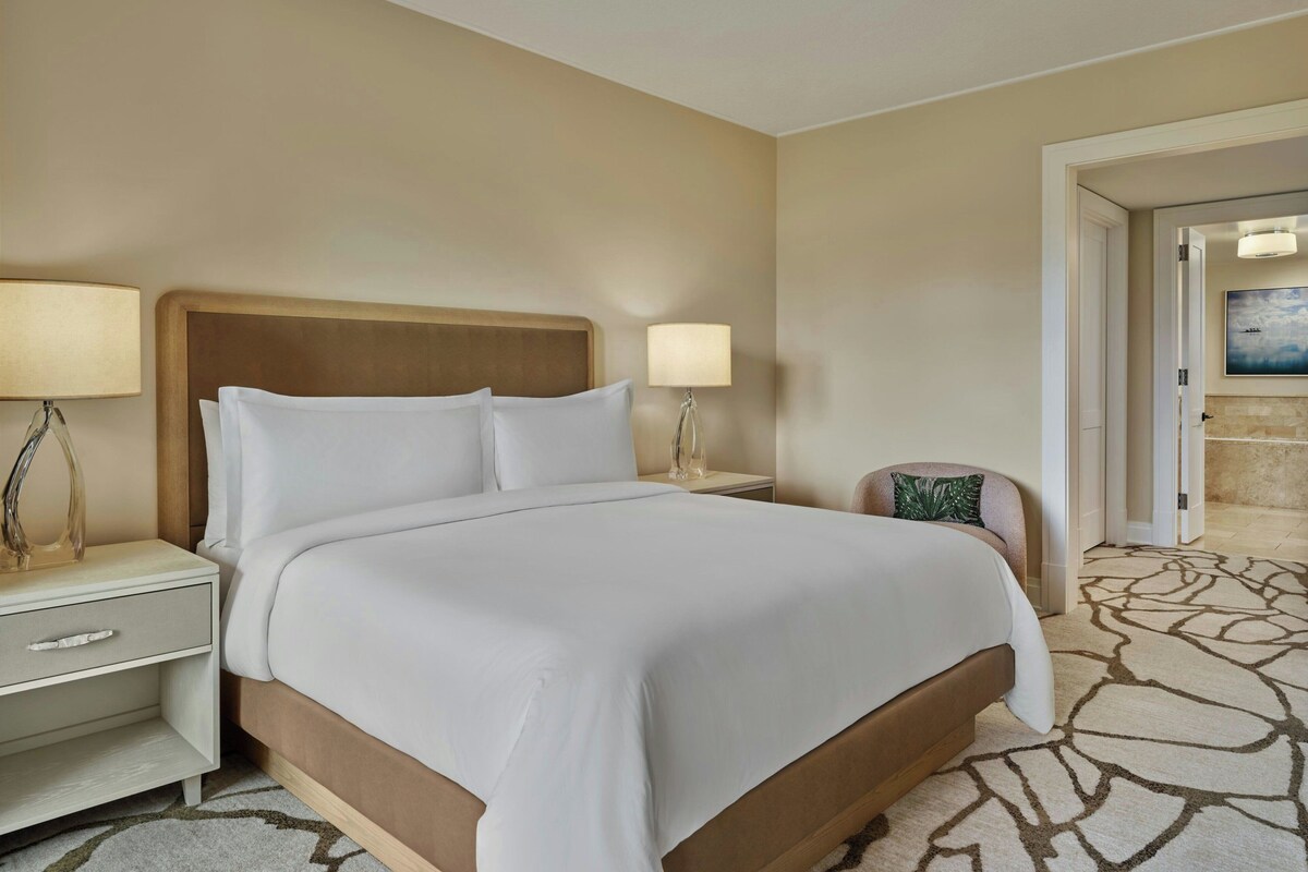 Een hotelkamer met een bed, twee nachtkastjes, een stoel en een deur naar een andere kamer.